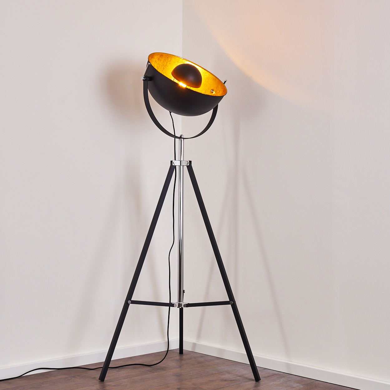 Eine schwarz-goldene Stehlampe mit Dreibein steht in einer Ecke. Die Lampe hat ein Scheinwerfer-Design und strahlt Licht aus.
