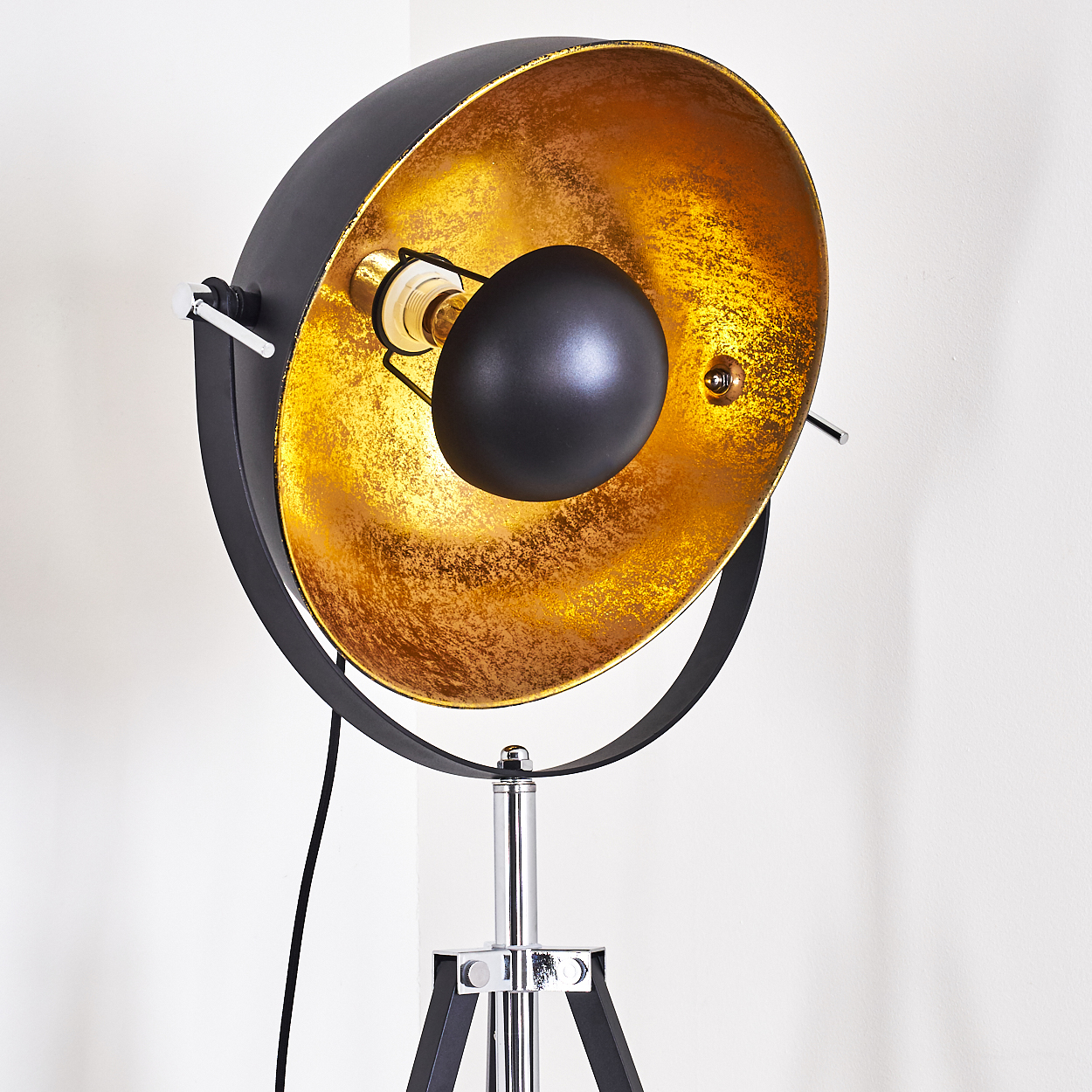 Eine moderne, schwarz-goldene Stehlampe vor einer weißen Wand. Die Lampe hat einen großen, schalenförmigen Schirm und ein Stativ.
