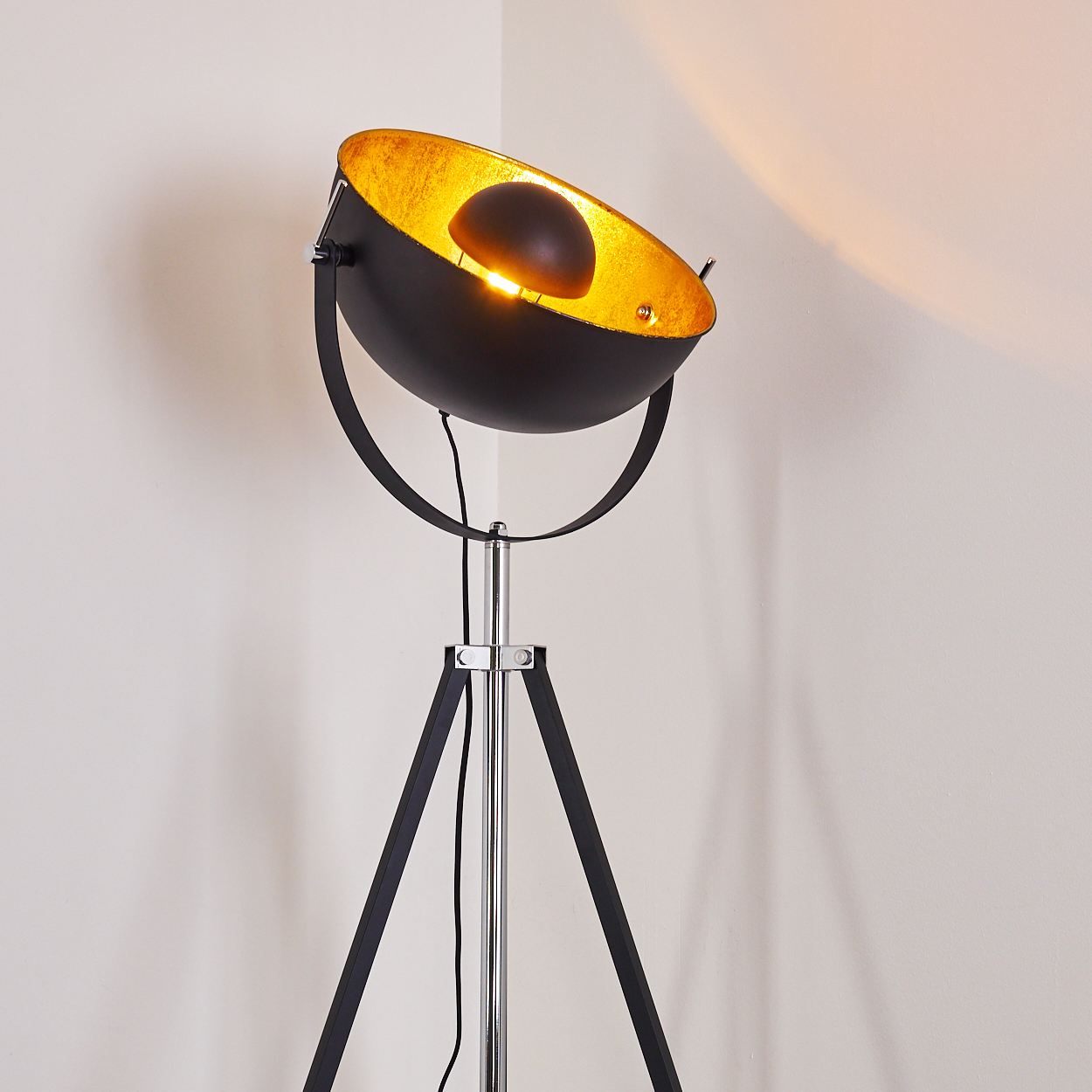 Eine schwarz-goldene Stehlampe steht in einer Ecke. Die Lampe hat ein Stativ, eine silberne Stange und eine Lichtschale.