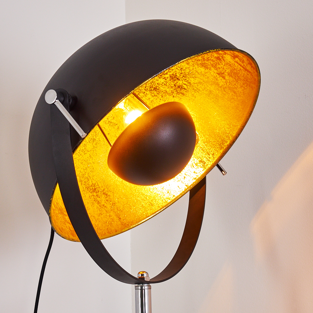 Eine moderne schwarze Stehlampe mit goldenem Innenraum. Die Lampe hat einen runden Schirm und einen Chromständer. Die Wand ist hellbeige.