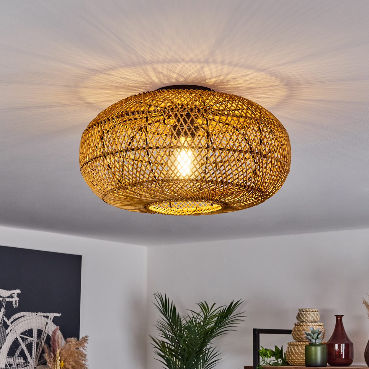 Eine gewebte Deckenlampe aus Rattan mit einer leuchtenden Glühbirne hängt. Schatten werden an die weiße Decke geworfen. Pflanzen und Dekoration sind im Hintergrund.