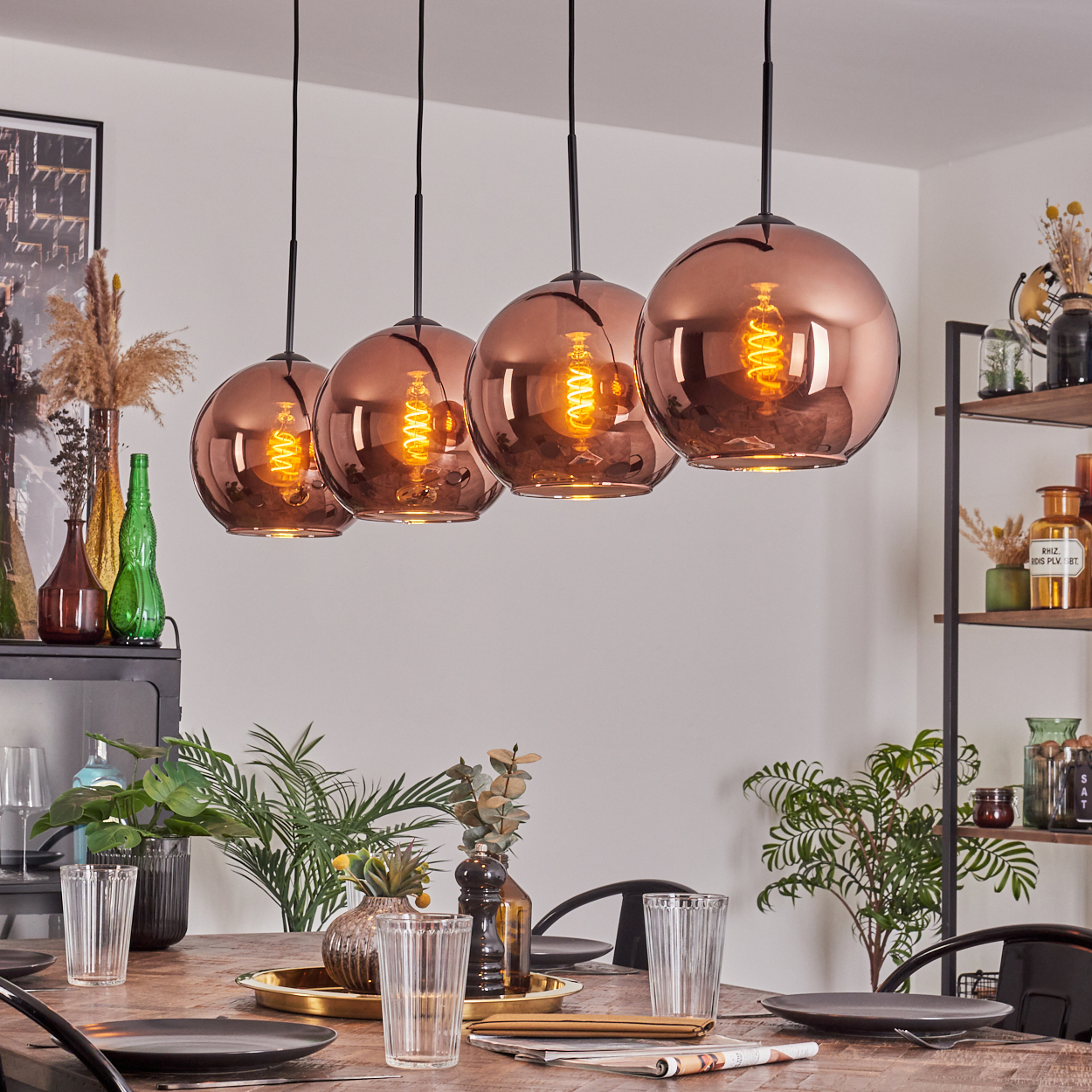 Esszimmer mit kupferfarbenen Pendelleuchten über einem Holztisch. Pflanzen, Gläser und ein Regal sind sichtbar.