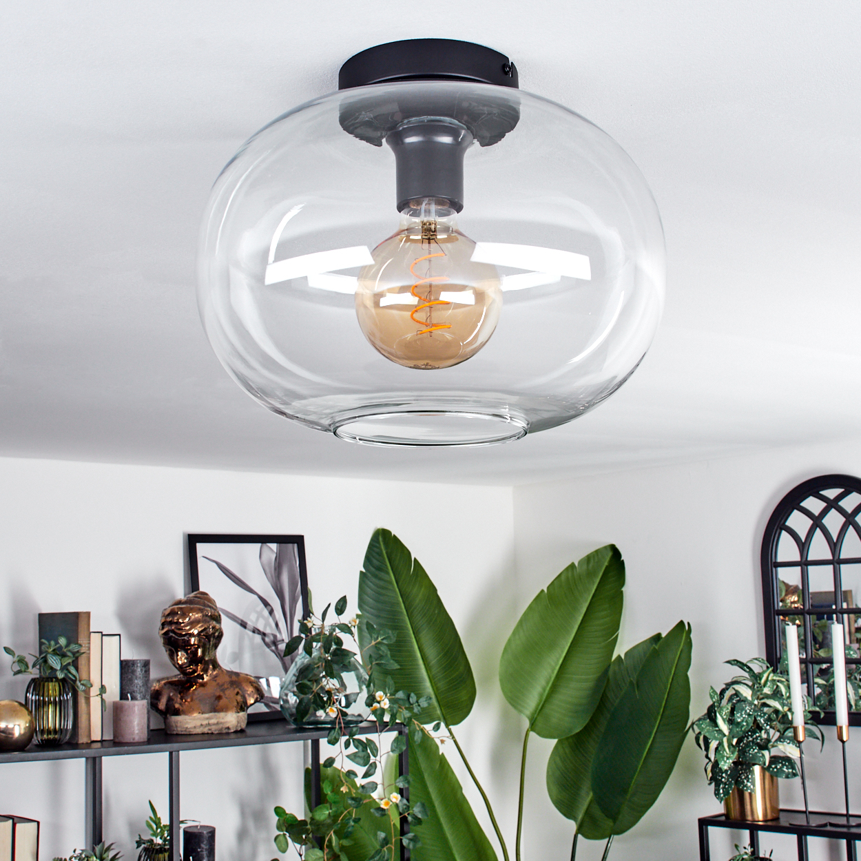Eine klare Glas-Deckenlampe mit sichtbarer Glühbirne. Darunter ist ein Regal mit Büste, Pflanzen und Kerzen zu sehen.