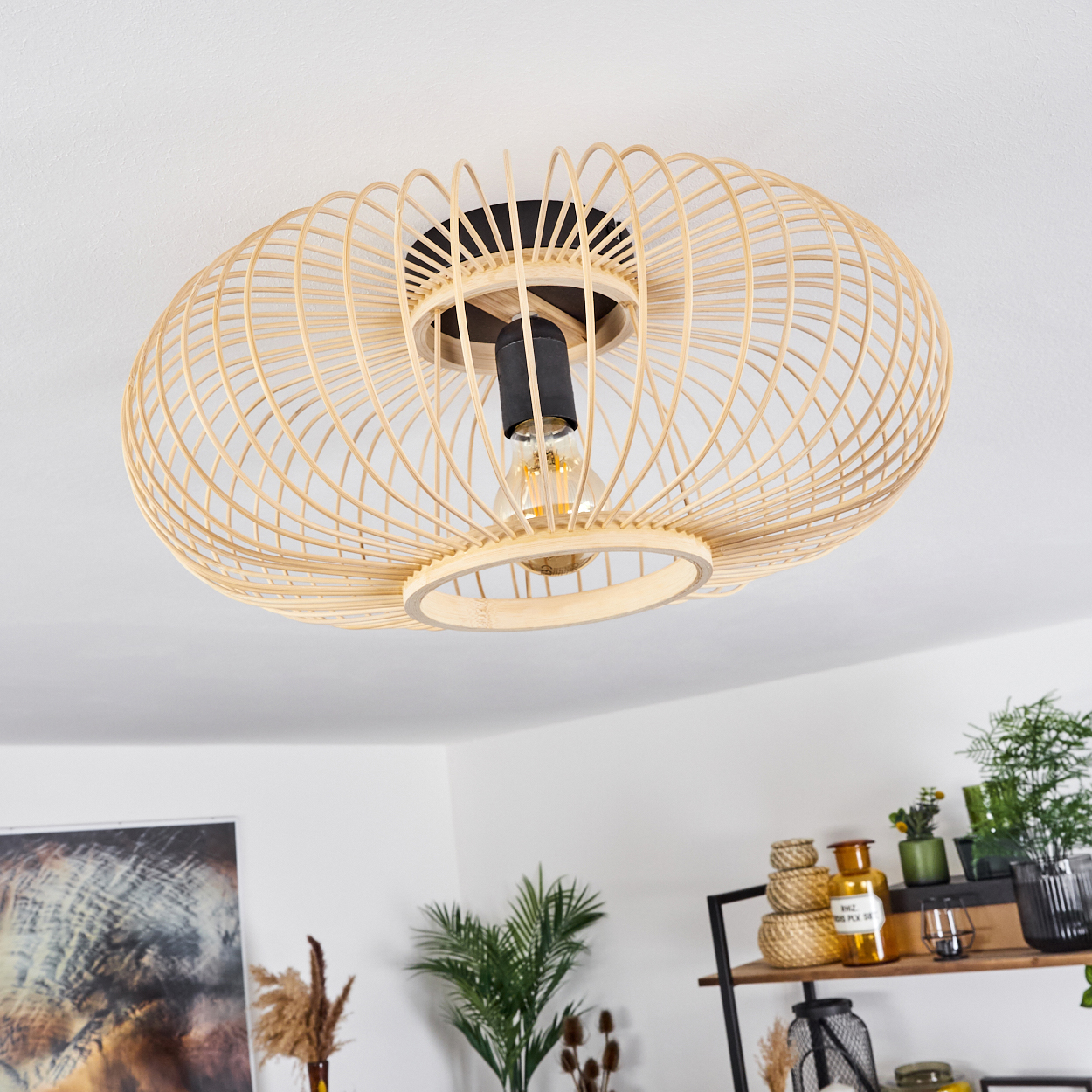 Eine gewebte, hellbraune, ovale Deckenlampe hängt. Darunter befinden sich Regale mit Vasen, Pflanzen und Dekorationsartikeln. Der Hintergrund ist weiß.