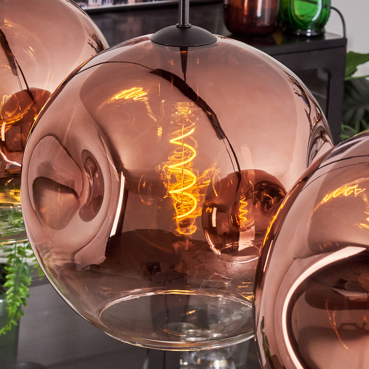 Nahaufnahme von kupferfarbenen Glas-Pendelleuchten mit Wendelglühlampen. Hintergrund mit Pflanzen und dunklen Regalen.