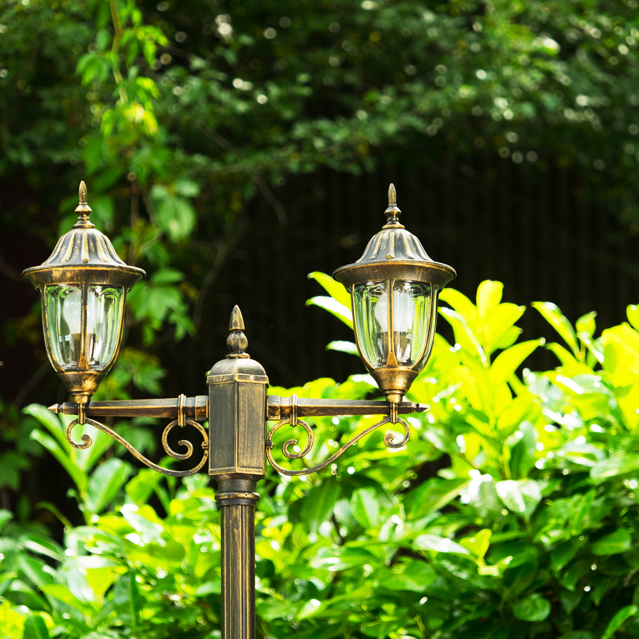 Ein dekorativer bronzefarbener Laternenpfahl mit zwei Lampen. Der Hintergrund ist üppiges grünes Laub.
