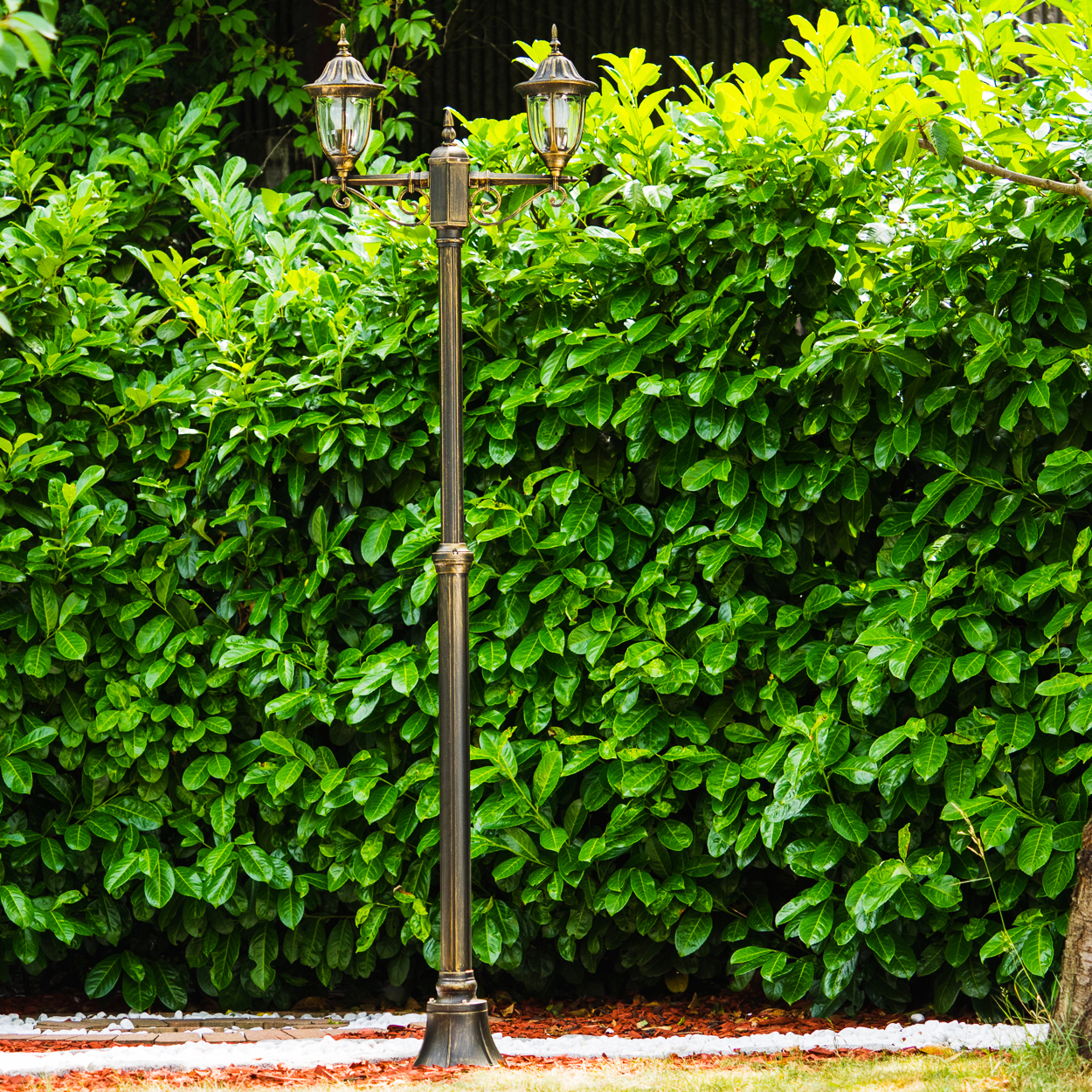 Eine bronzefarbene Laterne steht vor einer dichten, grünen Hecke. Die Lampe hat zwei Lichter. Weiße Steine und Gras befinden sich im Vordergrund.