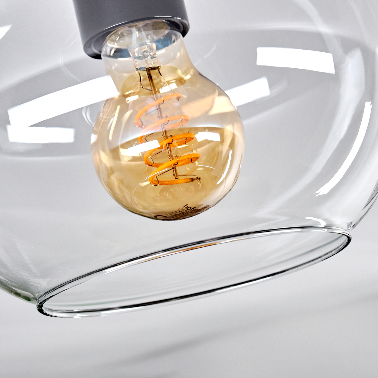 Nahaufnahme einer transparenten Glasleuchte mit einer großen, warmtonigen Glühbirne. Die Glühbirne ist von einem transparenten Glasschirm umgeben.