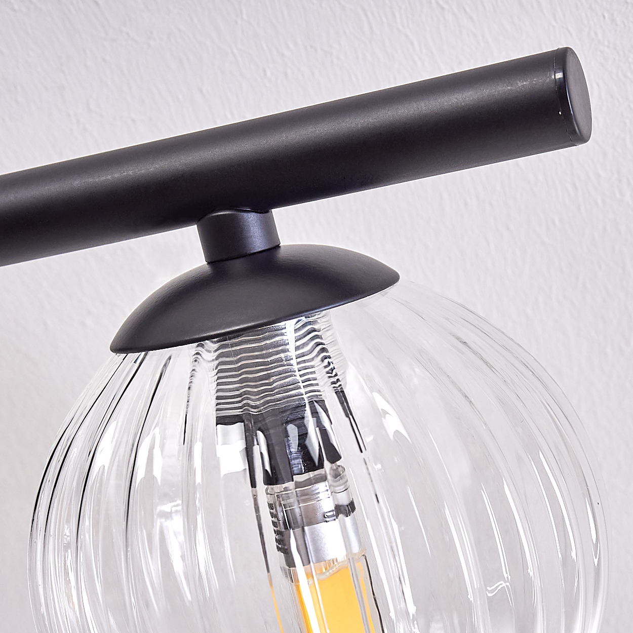 Nahaufnahme einer schwarzen Leuchte mit gerippter Glaskugel. Der Hintergrund ist eine schlichte weiße Wand, und das Licht ist an.