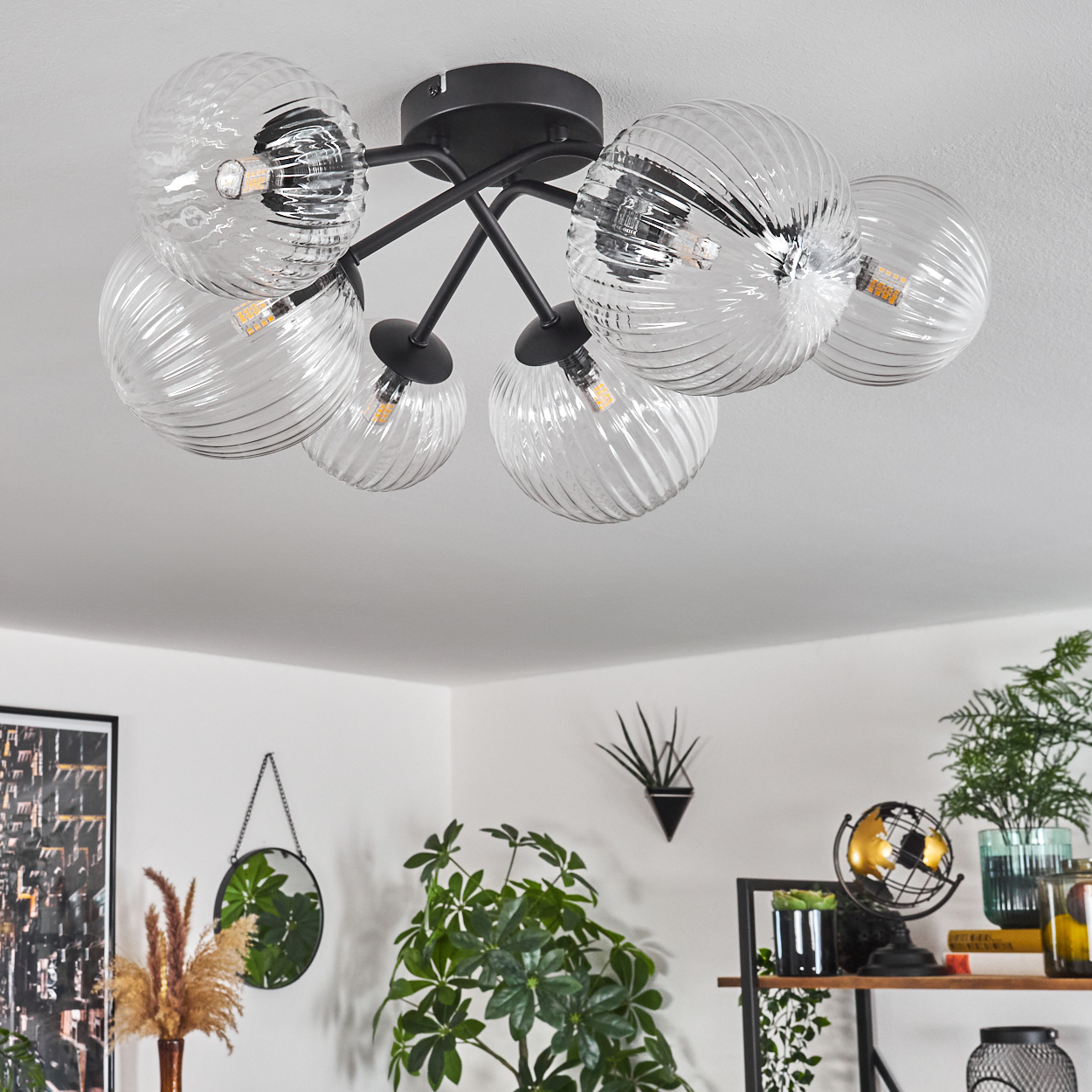 Eine moderne Deckenlampe mit sechs klaren Glaskugelschirmen. Der Raum hat Pflanzen, einen Spiegel und ein Regal mit Dekorationsartikeln.