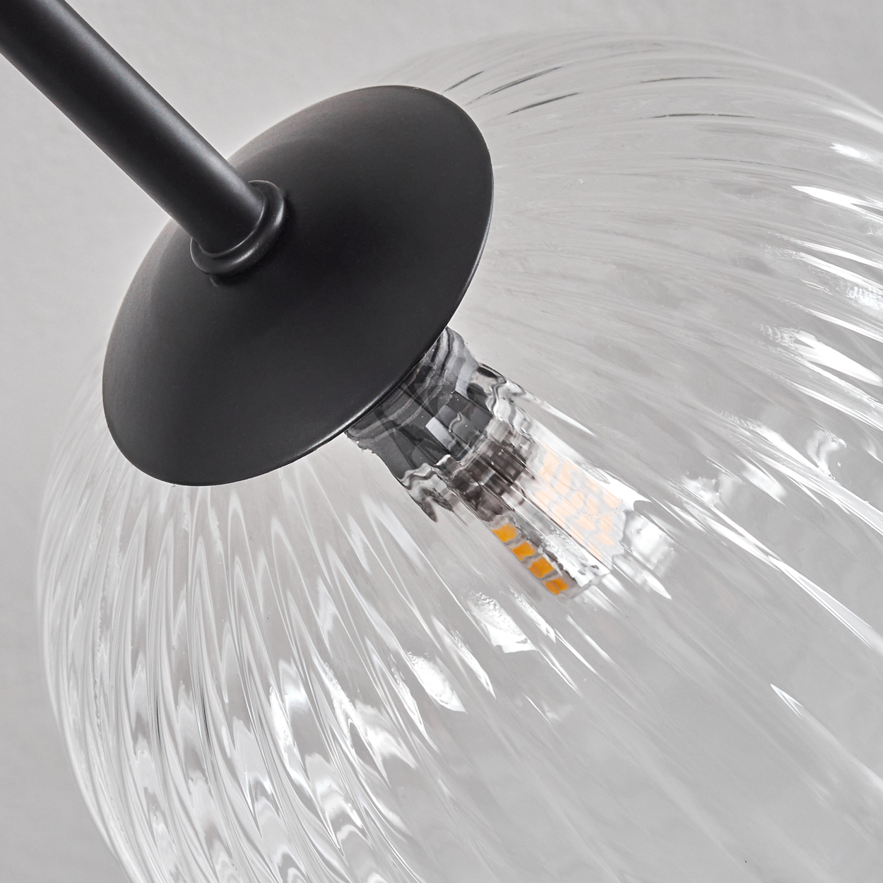 Nahaufnahme einer Glasleuchte. Ein schwarzer Metallarm und eine Platte tragen eine durchsichtige, strukturierte Glühbirne mit LED-Licht.