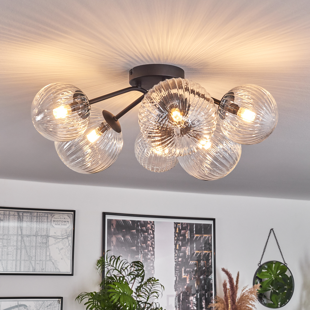 Eine moderne Deckenleuchte mit Glaskugeln. Das Licht ist an und beleuchtet den Raum. Kunstwerke und Pflanzen sind im Hintergrund sichtbar.