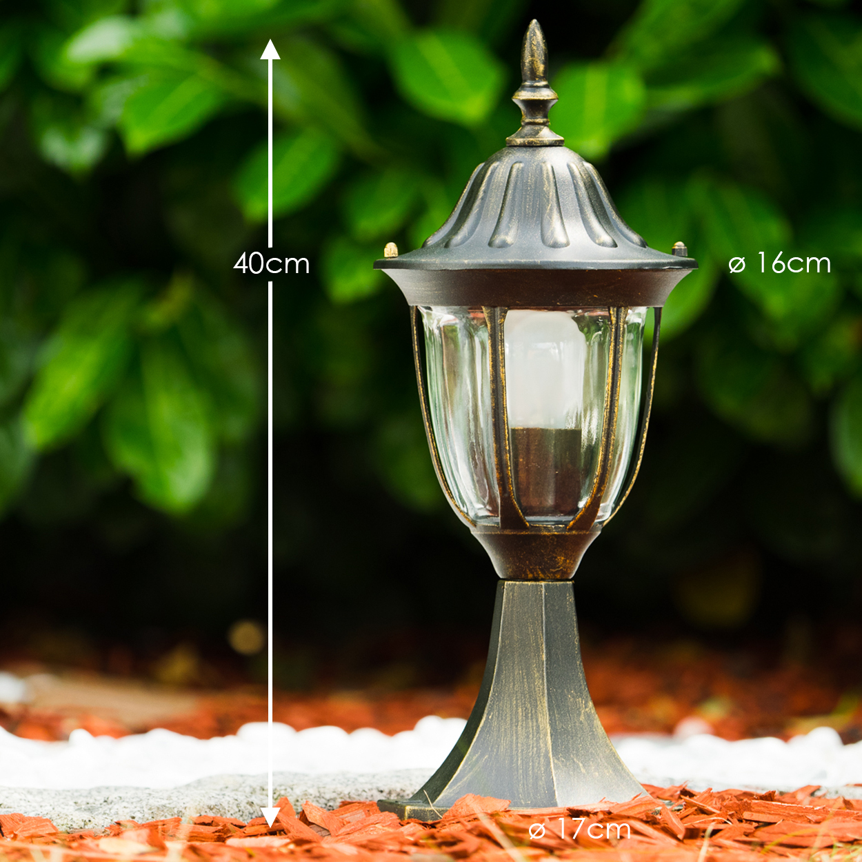 Eine bronzefarbene Außenlampe mit Glasgehäuse. Sie ist 40 cm hoch und hat einen Durchmesser von 16 cm. Die Basis ist 17 cm. Auf Mulch und Stein gesetzt.