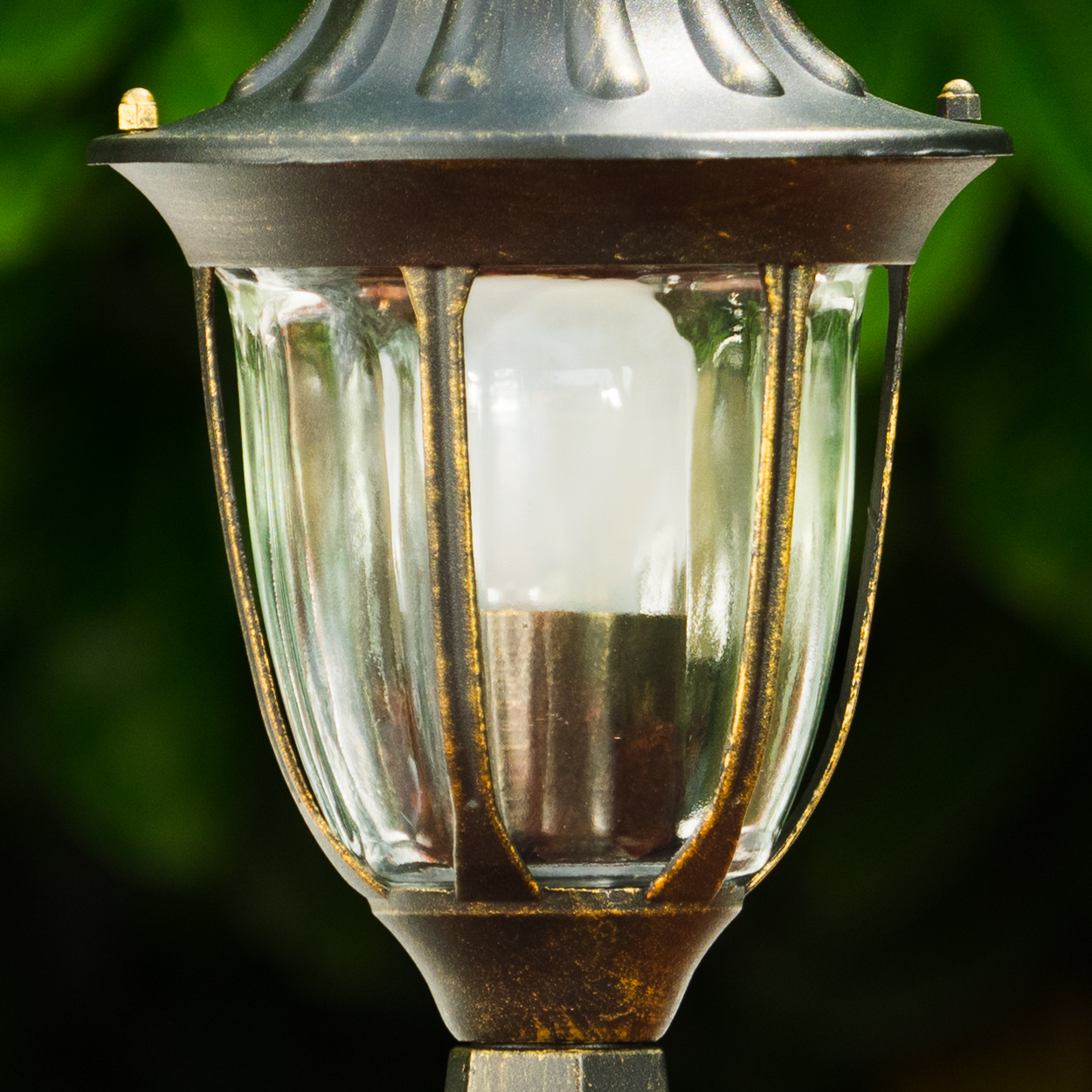 Nahaufnahme einer Vintage-Außenlampe mit Glaspaneelen und goldenem Rahmen. Der Hintergrund ist verschwommenes grünes Laub.