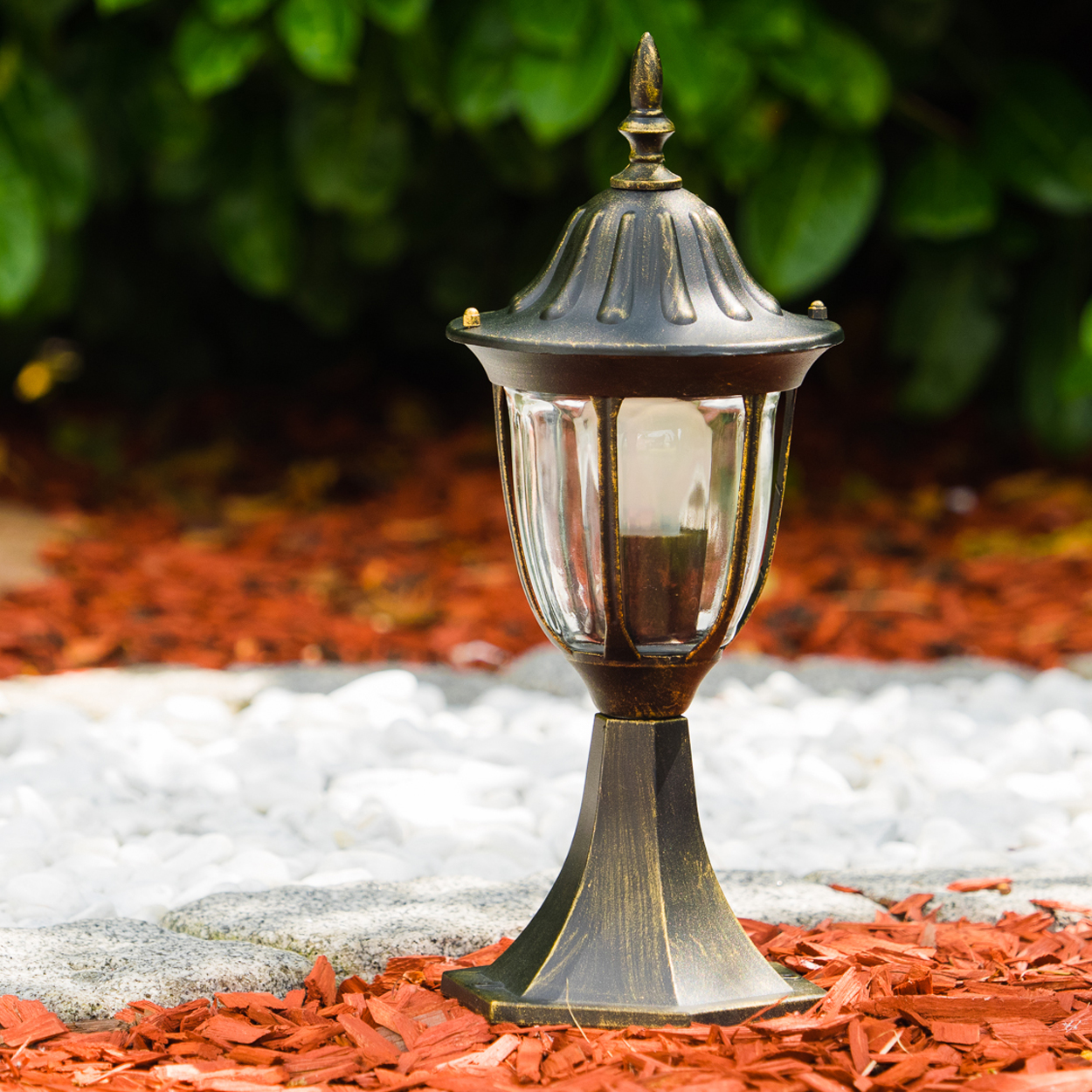 Eine dekorative Außenlampe mit Glasscheiben steht auf einem Bett aus rohem Mulch, weißen Steinen und einem Steinweg, mit grünem Laubwerk dahinter.