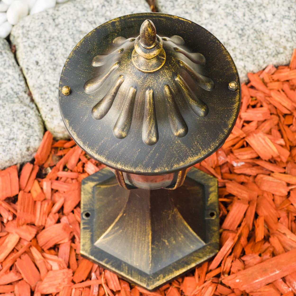 Eine bronzefarbene Außenlampe steht auf roter Mulch. Im Hintergrund sind graue Steine und weiße Kieselsteine.