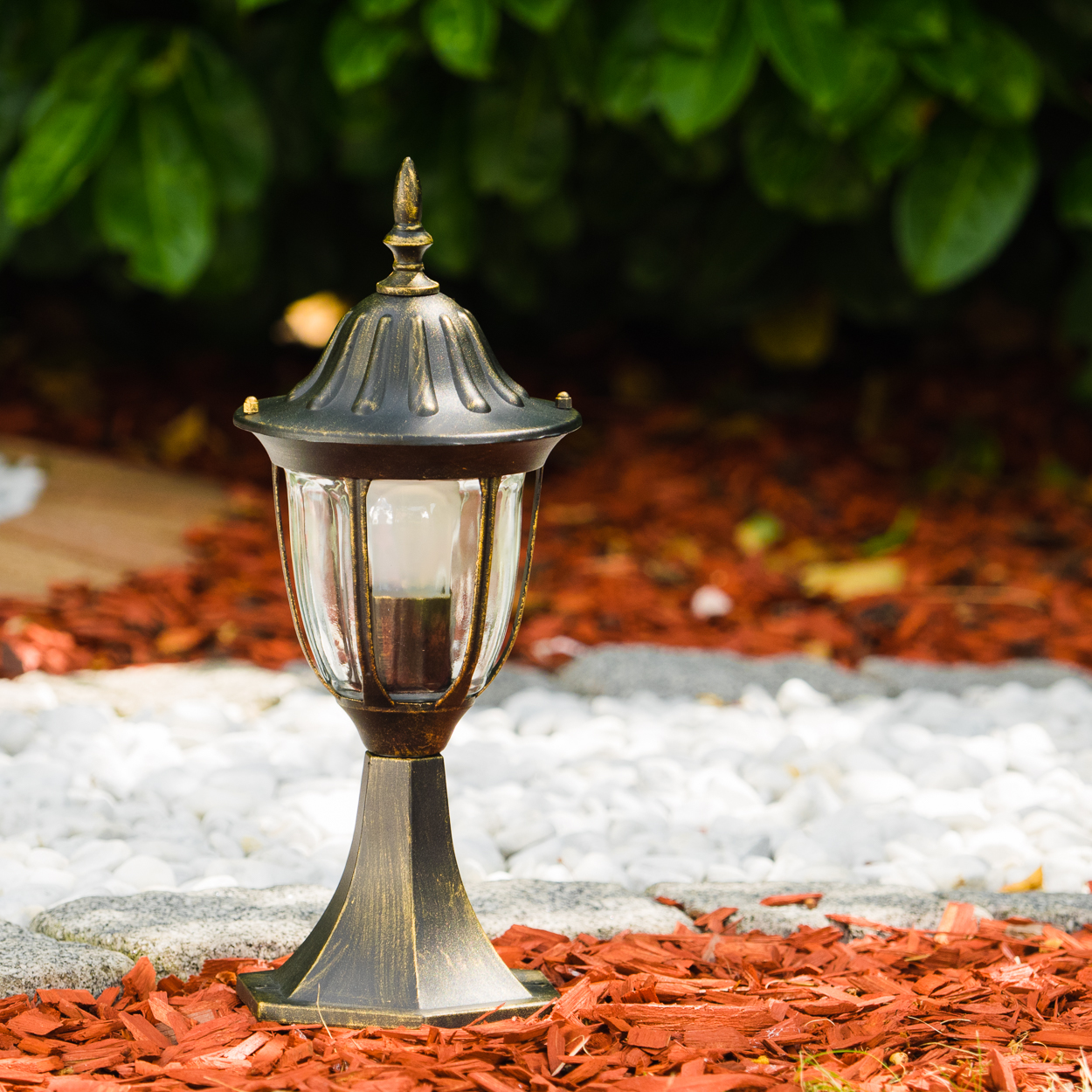 Eine verzierte Bronzelampe steht auf roter Mulch und weißen Steinen. Im Hintergrund sind grüne Büsche. Die Lampe hat ein klares Glas und eine Glühbirne.
