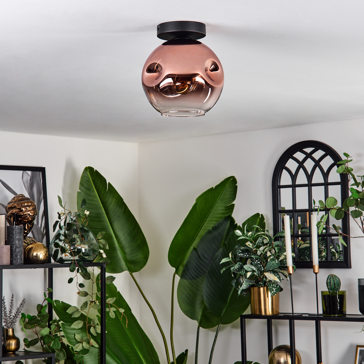 Ein modernes Wohnzimmer mit kupferfarbener Deckenlampe, Pflanzen, Regalen und einem dekorativen Spiegel.