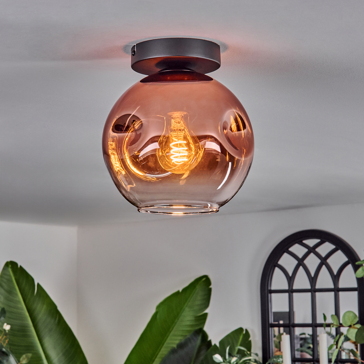 Eine moderne Deckenleuchte mit brauner Glaskugel. Das Licht ist an und beleuchtet den Raum. Pflanzen und ein Bogenfenster sind im Hintergrund.