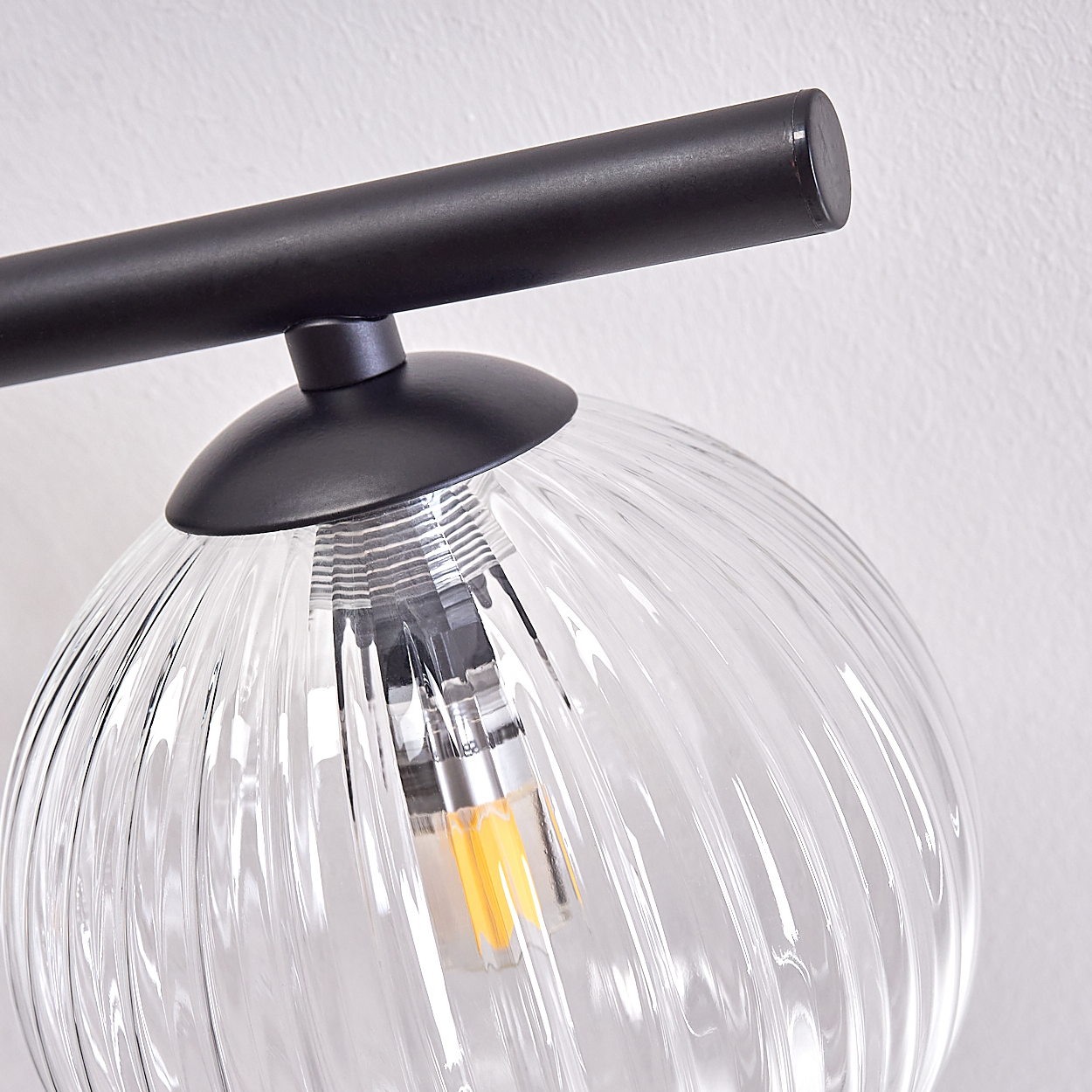 Nahaufnahme einer Lampe mit klarem, geripptem Glaskugel. Ein schwarzer Metallarm und Lampensockel sind vor einer weißen Wand sichtbar.