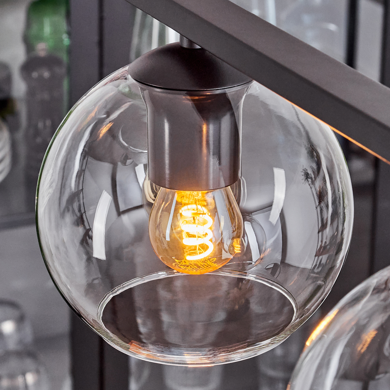Nahaufnahme einer modernen Leuchte. Ein runder, klarer Glasschirm umgibt eine leuchtende Glühbirne mit Spiralfilament. Dunkle Metallakzente.