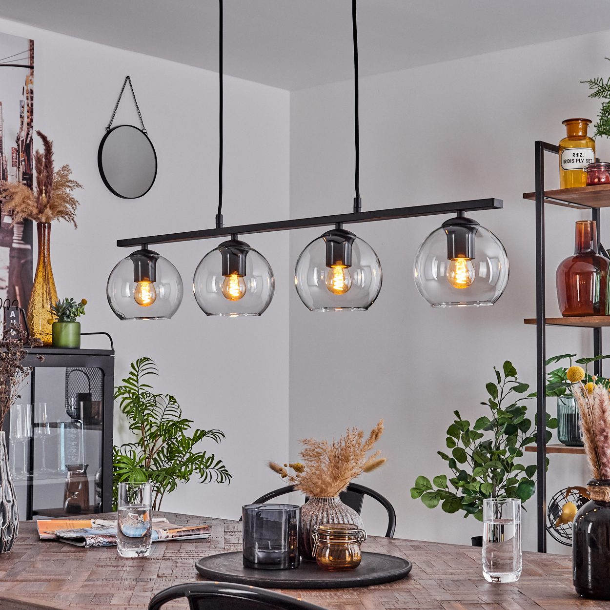 Esszimmer mit Holztisch, schwarzem Kronleuchter mit vier Glaskugeln, Pflanzen und Dekorationen.