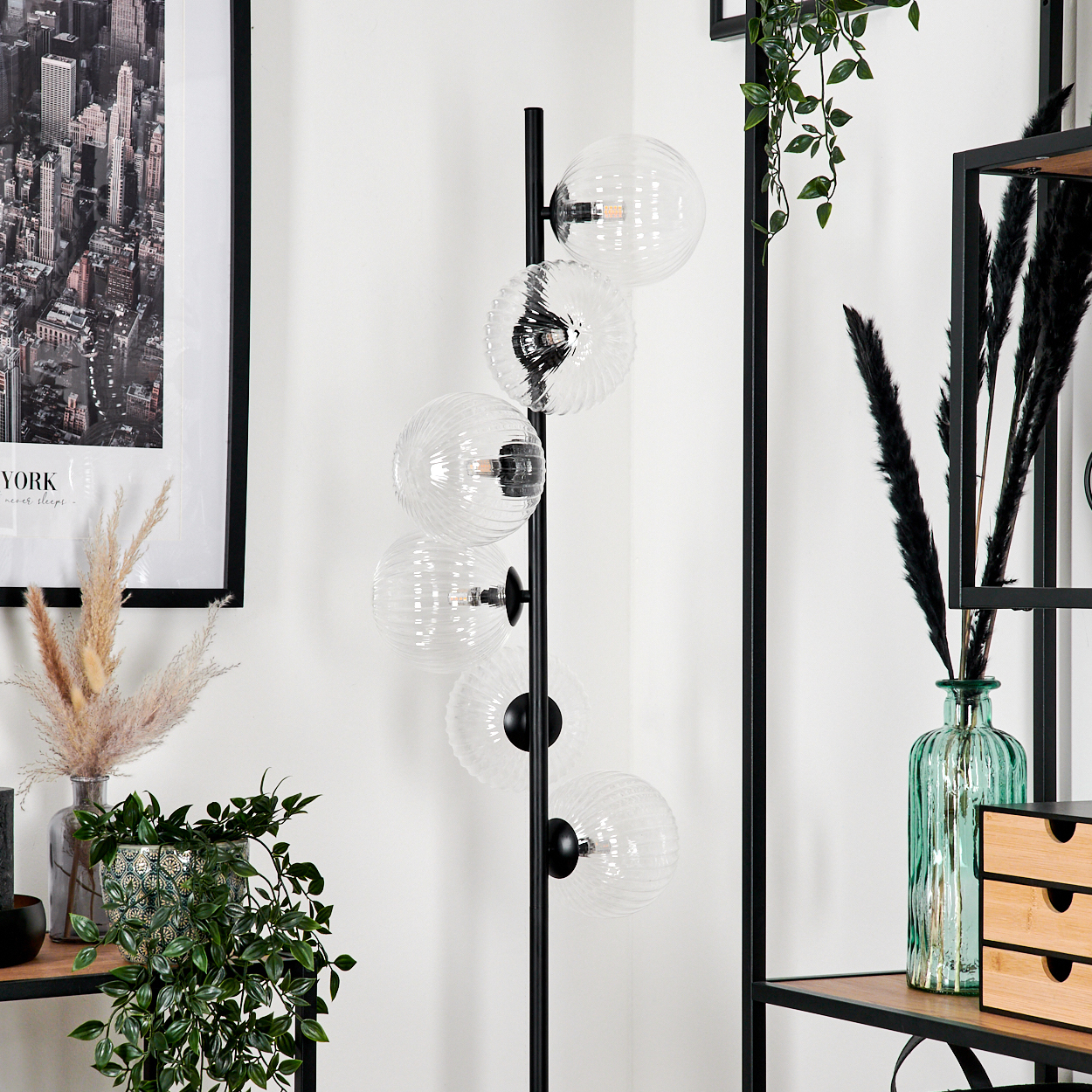 Ein modernes Zimmer mit einer schwarzen Stehlampe mit Glaskugeln. Es gibt Pflanzen, Regale und ein Stadtbildbild an der Wand.