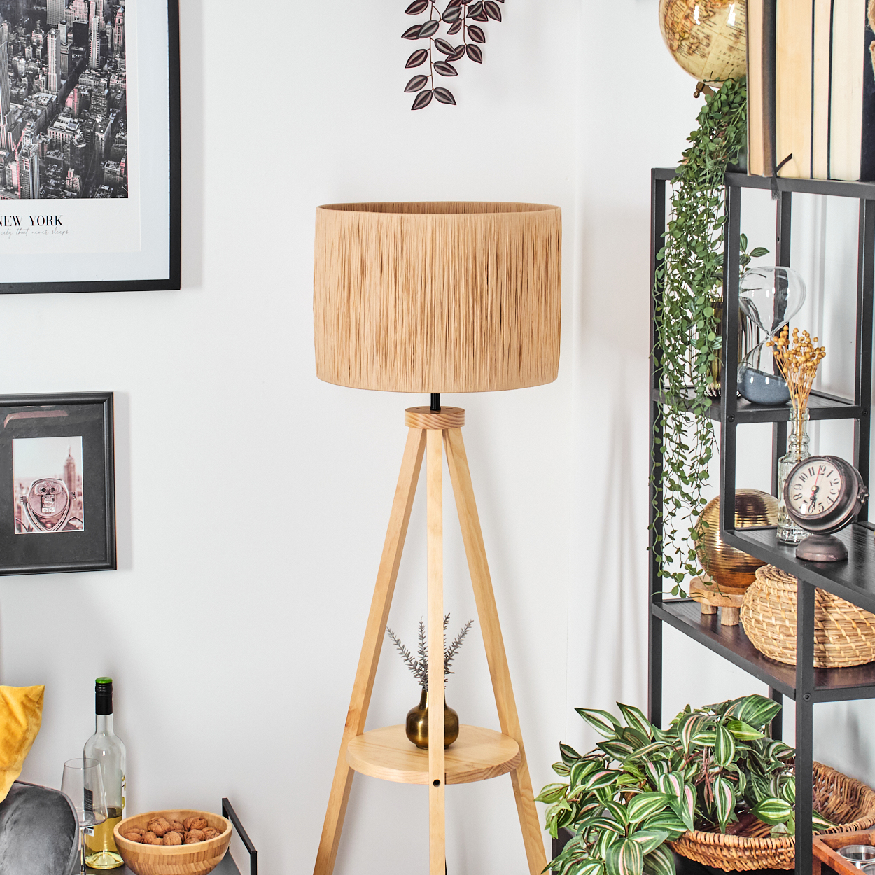 Eine Holz-Stativlampe mit beigem Schirm steht vor einer weißen Wand. Regale rechts halten Deko und Pflanzen.