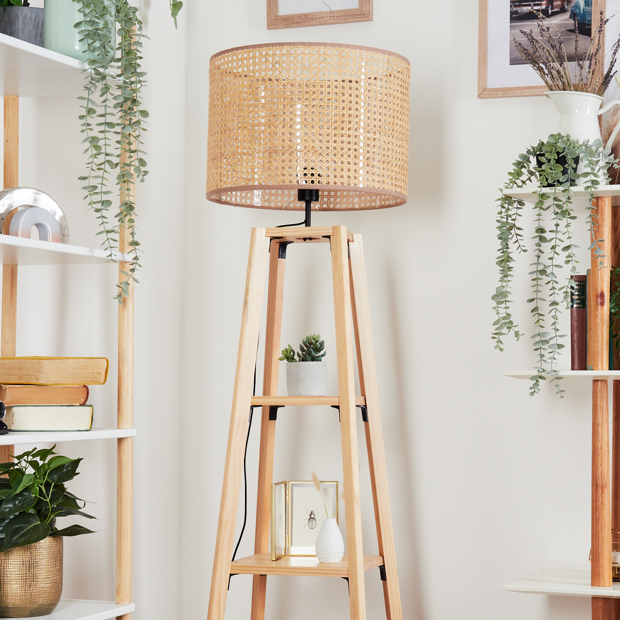 Eine gewebte Lampe steht auf einem Holzrahmen, Regale mit Pflanzen und Büchern sind an den Seiten. Der Hintergrund ist weiß.
