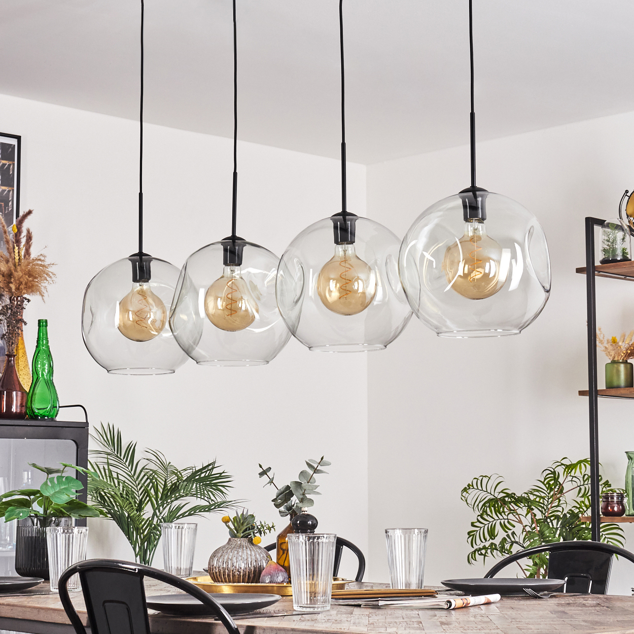Vier Glaskugel-Pendelleuchten hängen über einem Holztisch, der mit Tellern und Gläsern gedeckt ist. Pflanzen und Dekorationen sind sichtbar.