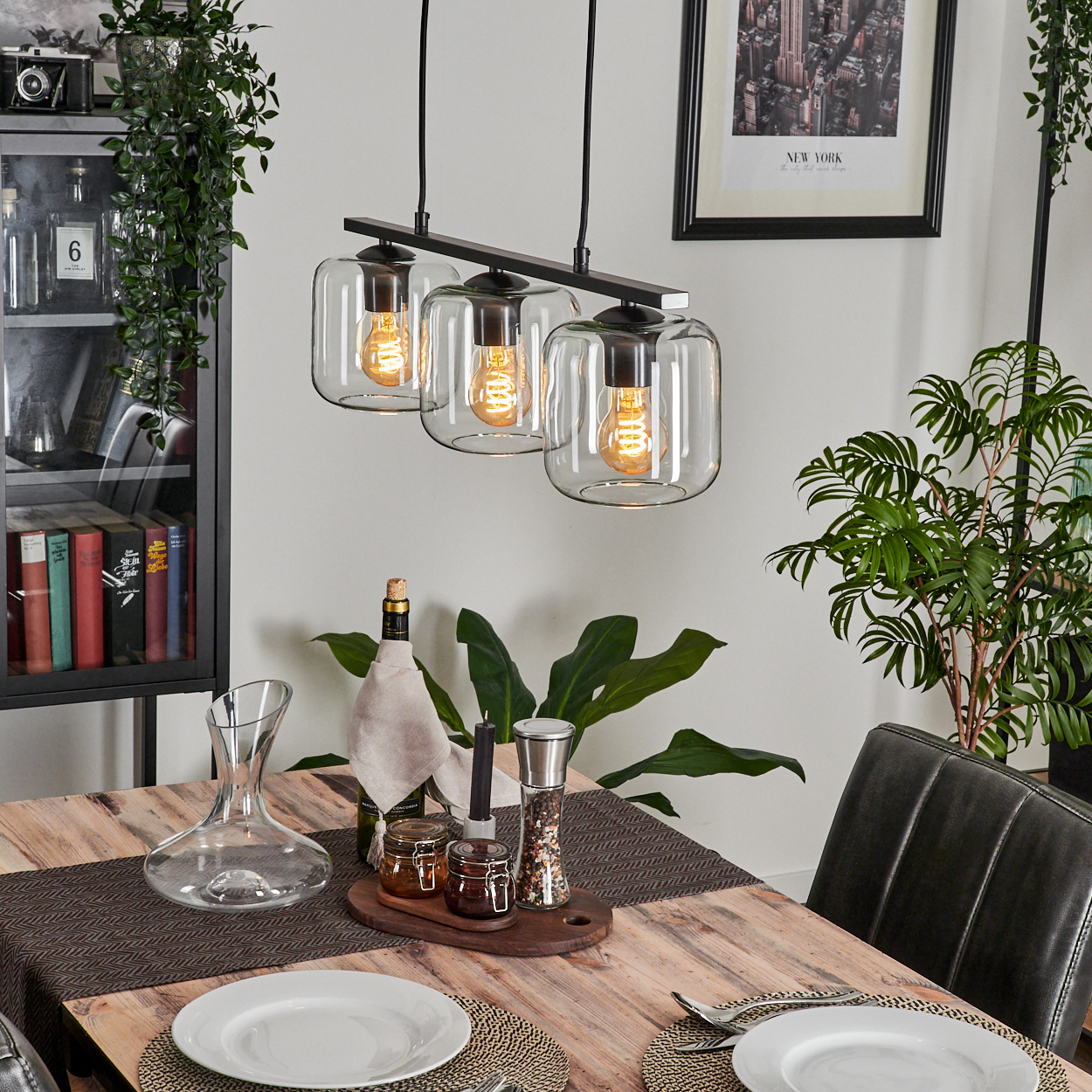 Esszimmer mit Holztisch, dreifacher Leuchte, Pflanzen und Schrank. Teller und Besteck sind gedeckt.
