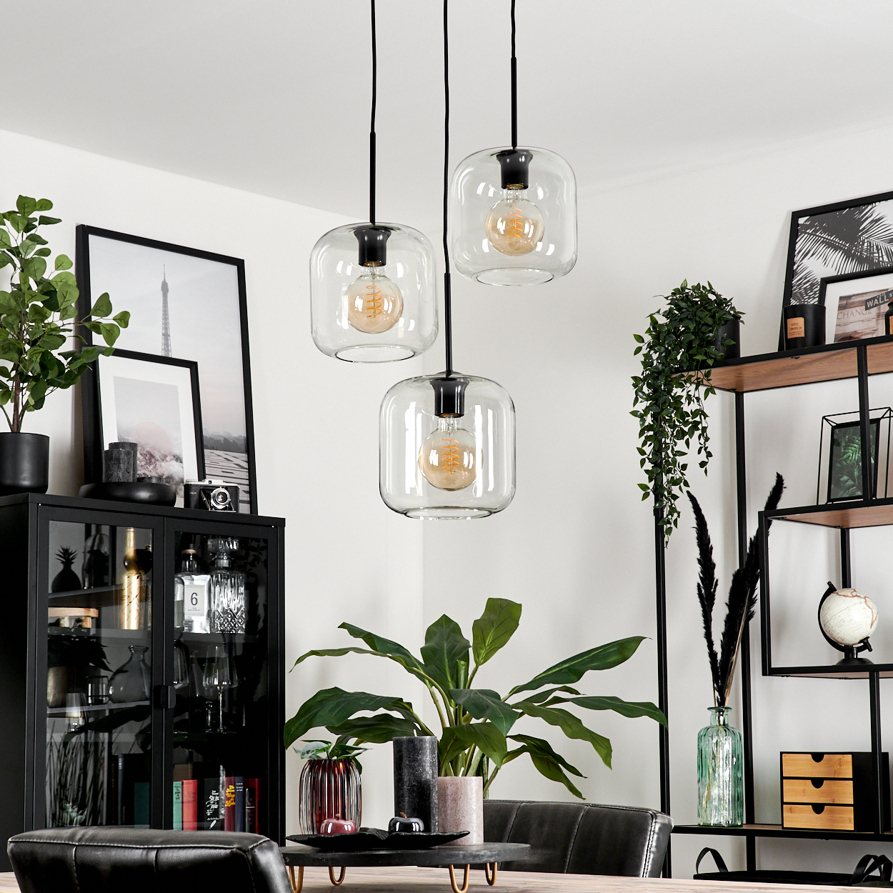 Ein Esszimmer mit drei Hängelampen. Ein schwarzer Schrank und Regal zeigen Pflanzen, Fotos und Dekorationsartikel. Darunter steht eine große Pflanze.