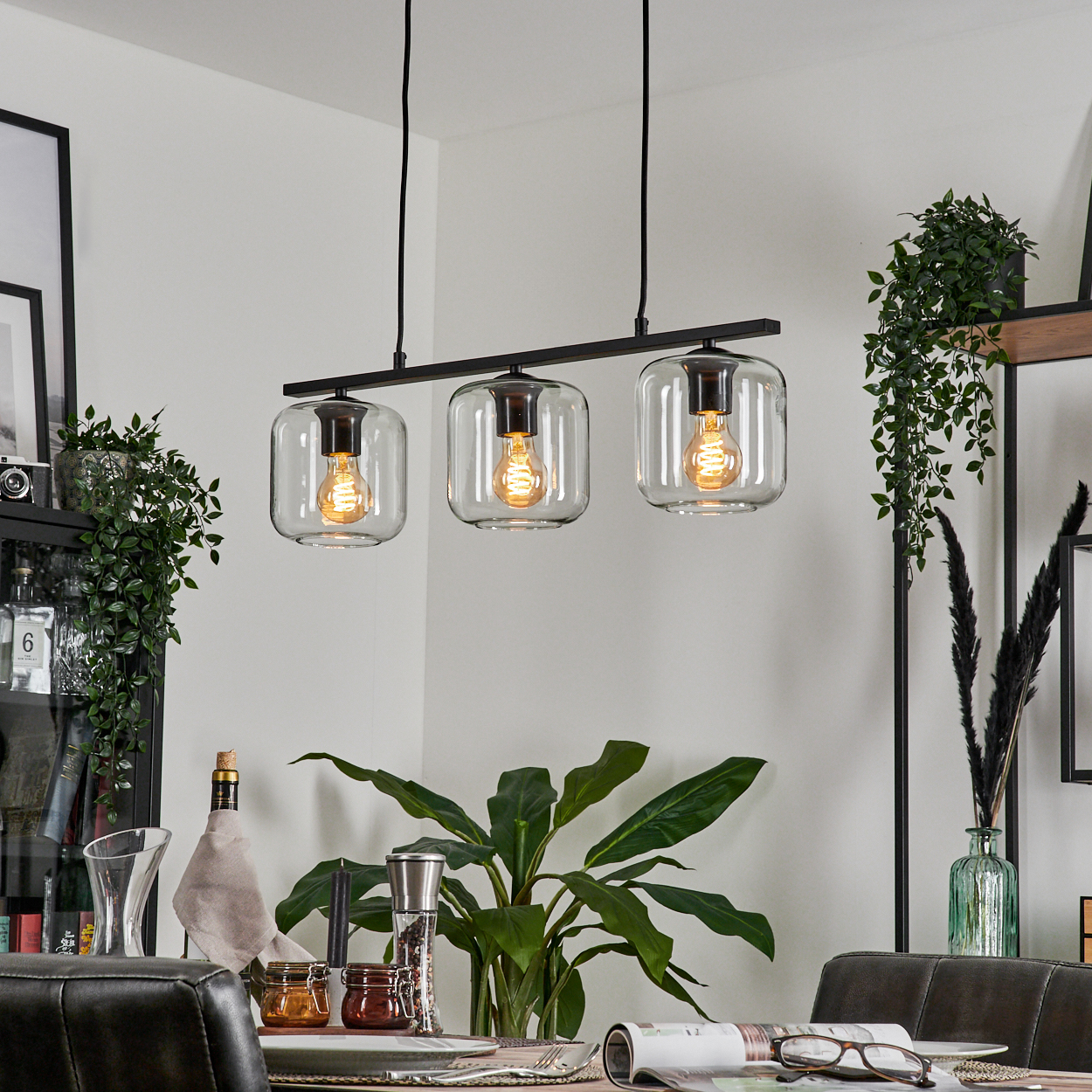 Esszimmer mit moderner Leuchte. Drei transparente Glasschirme mit Glühbirnen hängen über einem Tisch mit Pflanzen und Dekoration.