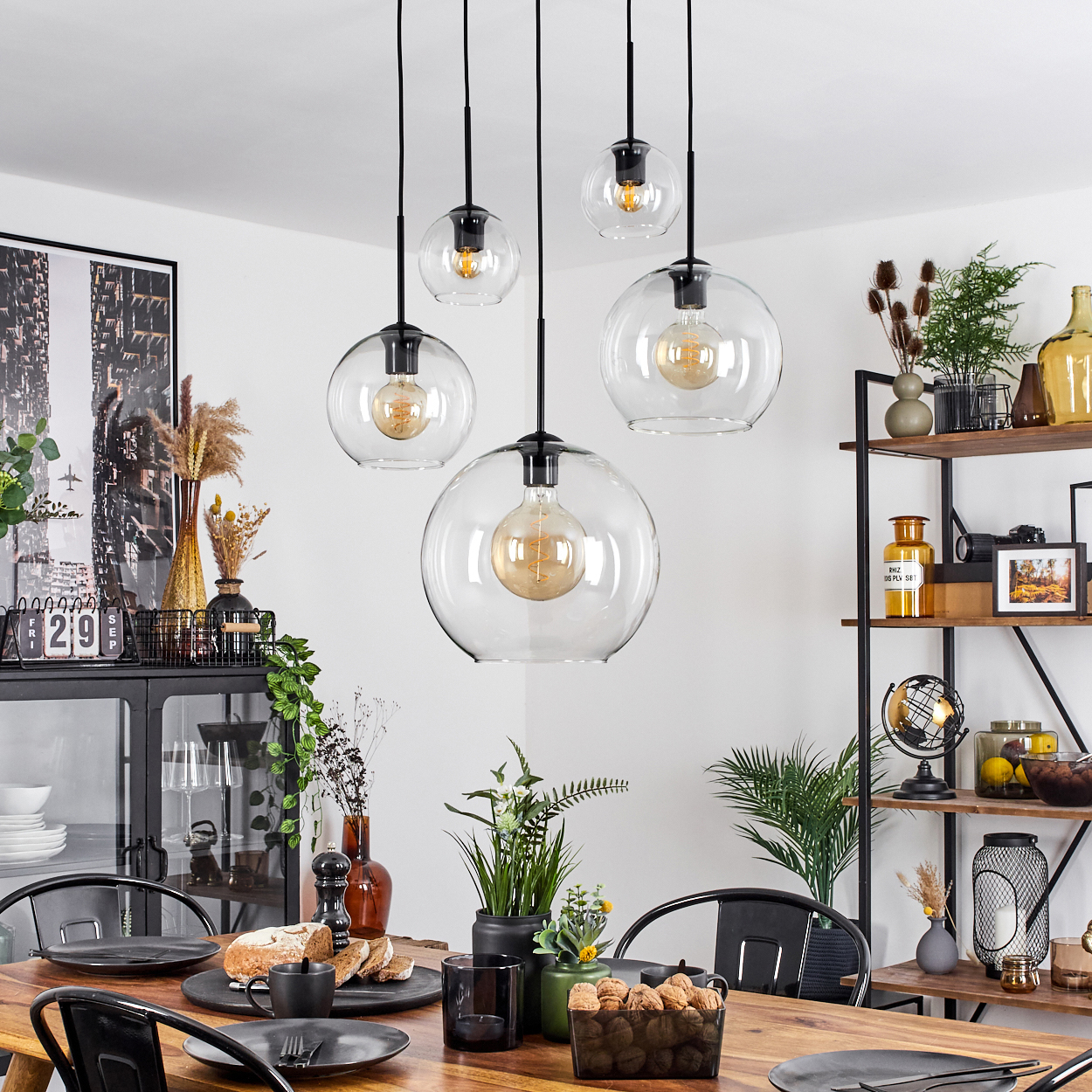 Esszimmer mit Holztisch. Glaskugel-Pendelleuchten hängen darüber. Teller, Brot und Dekorartikel schmücken den Tisch. Regale rechts.