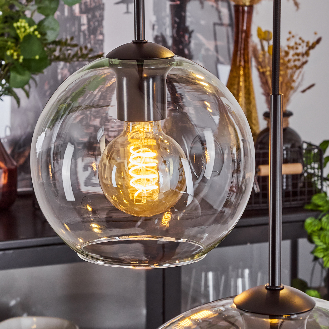 Eine klare Glaskugel-Hängelampe mit einer leuchtenden, spiralförmigen Glühbirne. Eine weitere Lampe und dekorative Vasen sind sichtbar.