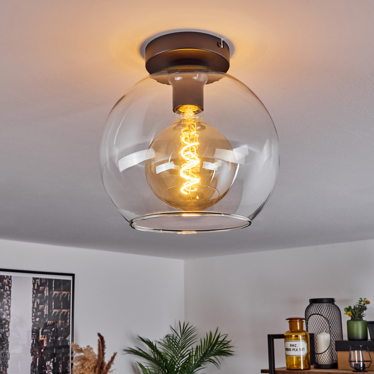 Eine klare Deckenleuchte mit Glaskugel und einer Glühbirne mit Spiralfaden. Die Lampe ist an einer weißen Decke montiert, darunter ein Regal.