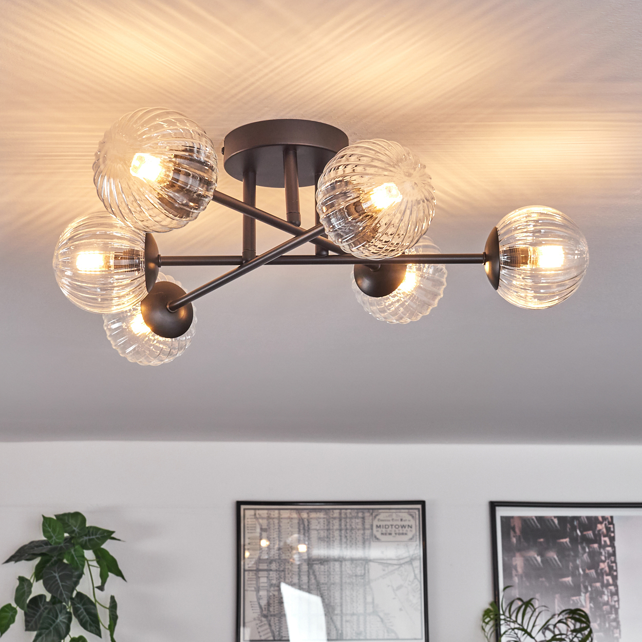 Deckenleuchte mit klaren Glaskugeln. Braune Metallarme und -sockel. Weiße Decke. Kunstwerke und Pflanzen im Hintergrund.