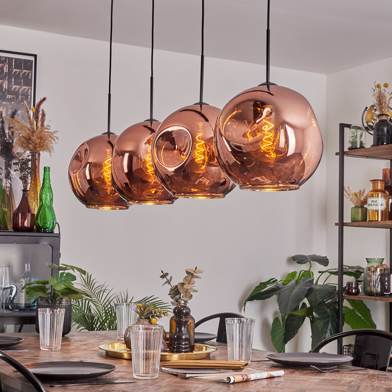 Vier kupferfarbene Pendelleuchten hängen über einem Holztisch, der für eine Mahlzeit gedeckt ist. Pflanzen und Dekorationsartikel sind im Hintergrund.