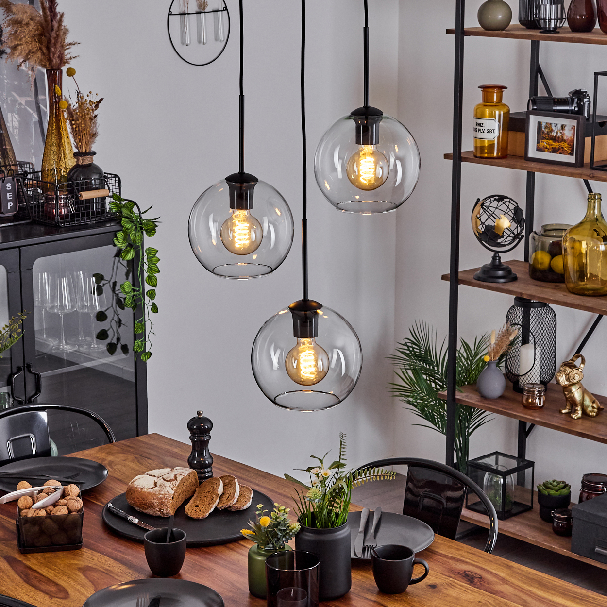 Esszimmer mit Holztisch, Glaskugel-Pendelleuchten, Brot, Tellern und Dekorationsartikeln auf Regalen.