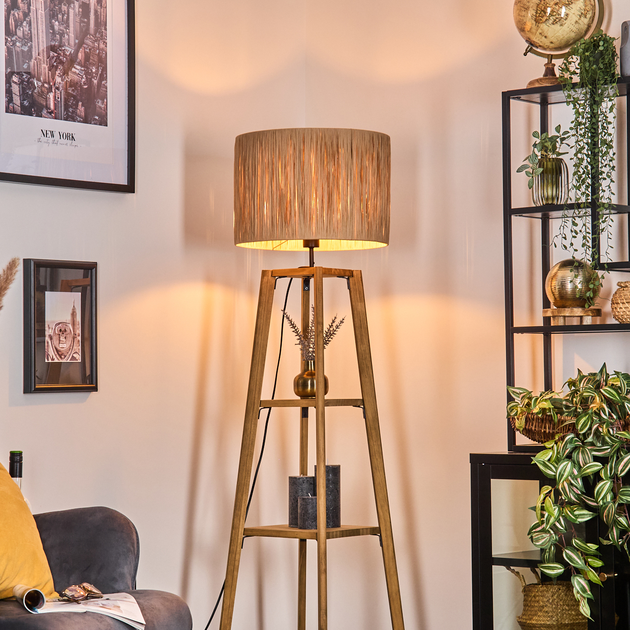 Eine hölzerne Stehlampe mit beigem Lampenschirm steht in einem Raum. Regale und Bilder sind im Hintergrund.
