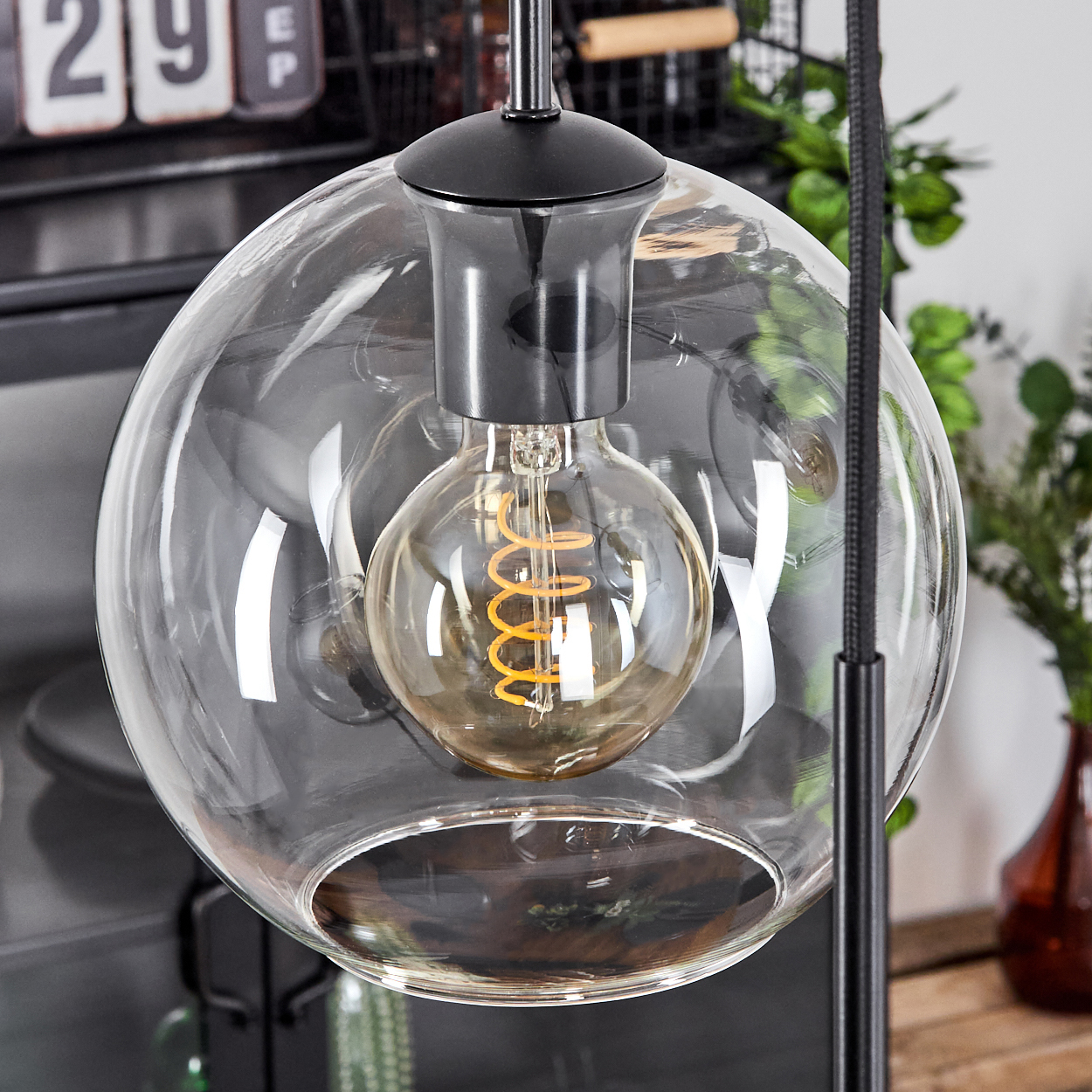 Eine klare Glaskugel-Pendelleuchte mit einer leuchtenden Glühbirne mit Spiralfaden. Dahinter ein verschwommener Hintergrund mit Objekten und Pflanzen.