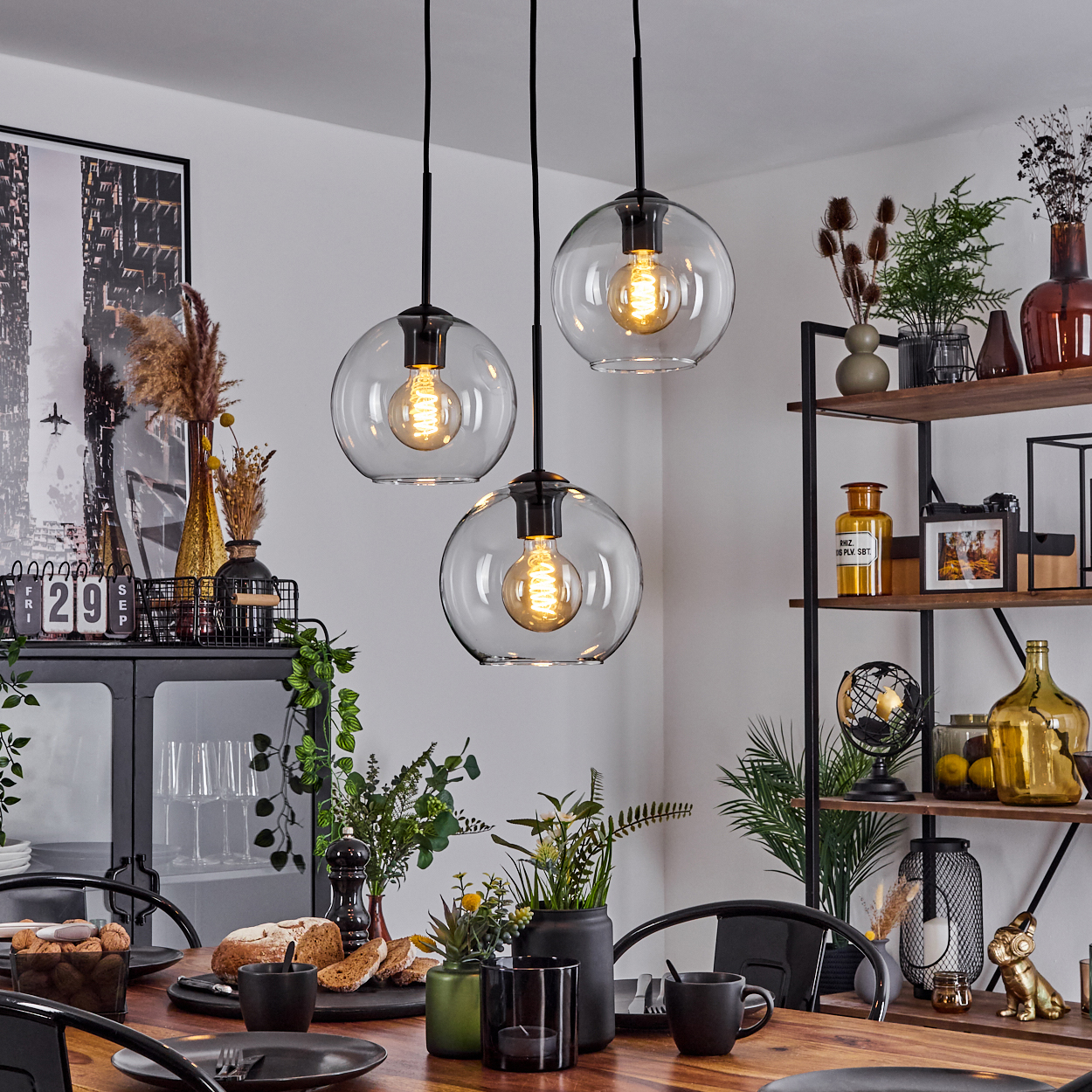 Esszimmer mit Holztisch, Glaslampen und dekorativen Elementen. Brot und Pflanzen auf dem Tisch, Regale im Hintergrund.