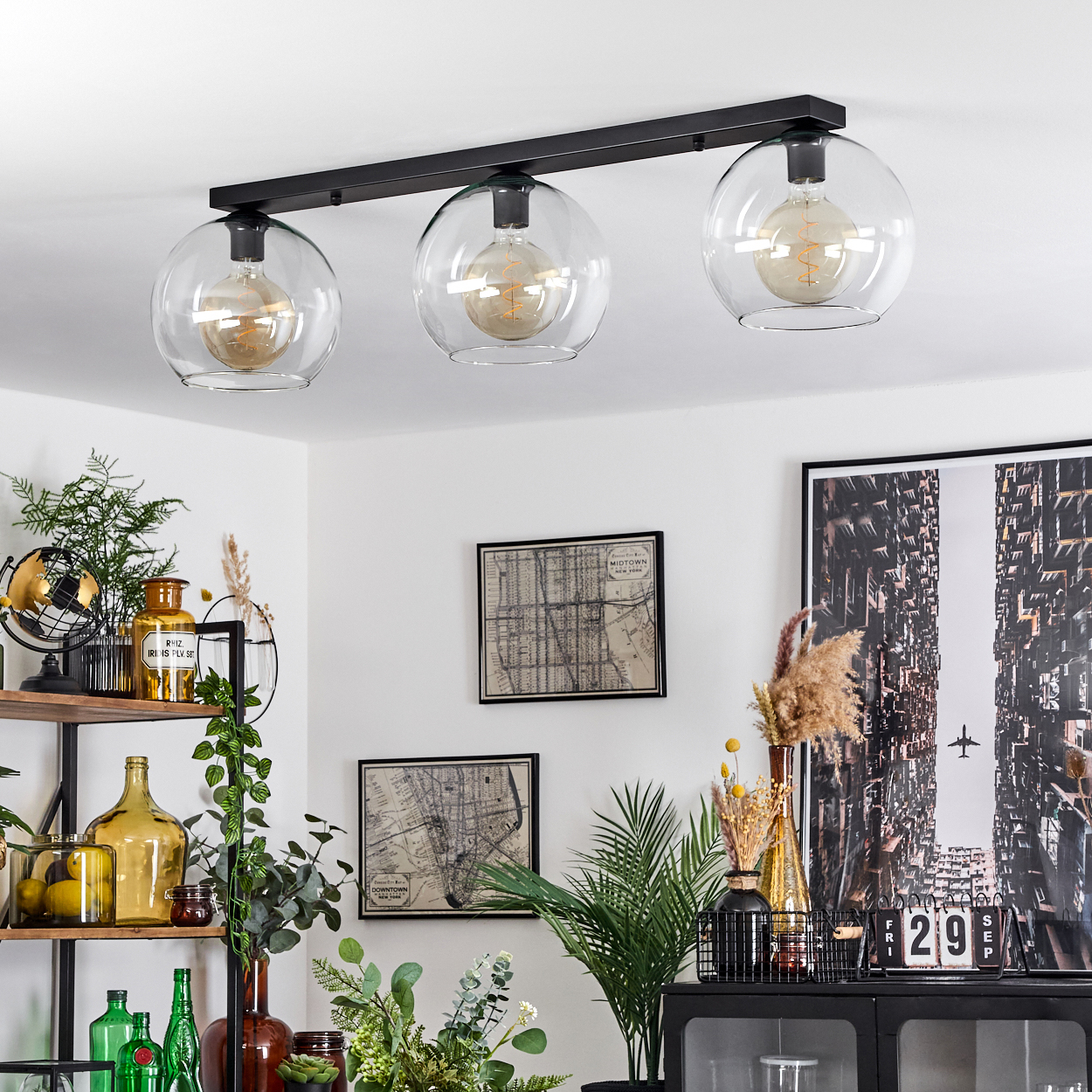 Deckenlampe mit drei Glühbirnen. Regale mit Dekoration und gerahmten Karten. Schwarzer Schrank mit Kalender. Pflanzen und ein großes Bild mit Flugzeug.