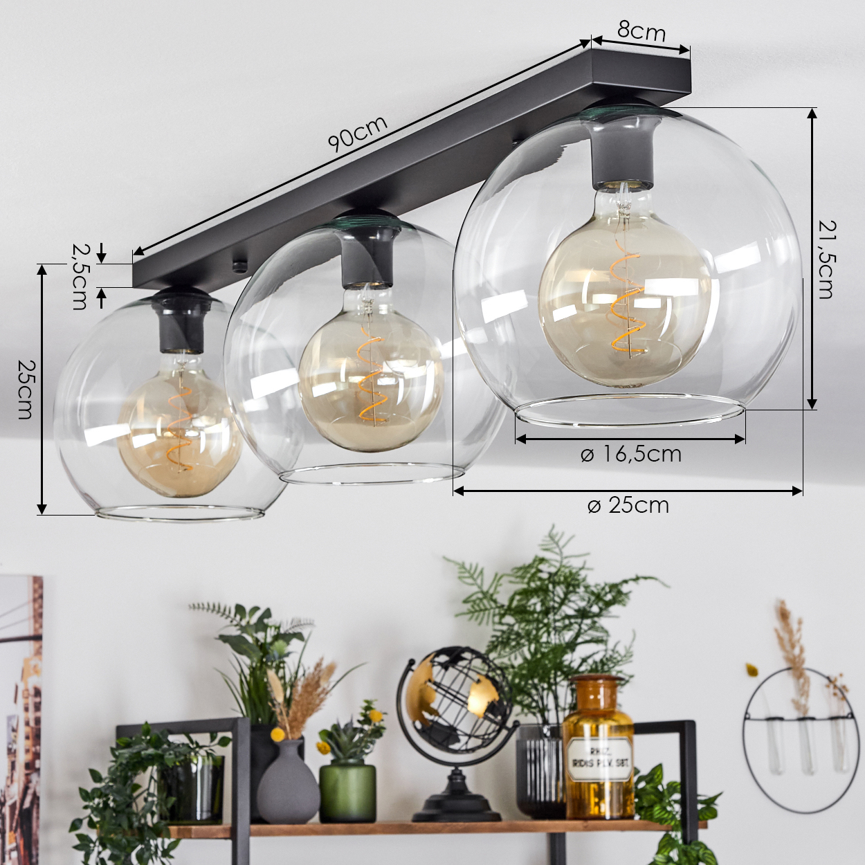 Deckenleuchte mit drei Glühbirnen und transparenten Kugeln, schwarze Halterung. Darunter ein Regal mit Pflanzen, einem Globus und Dekorationsartikeln.