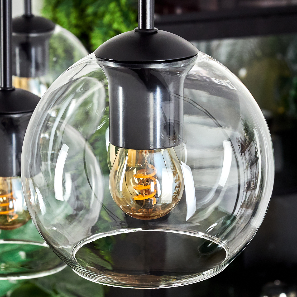 Nahaufnahme einer Glaskugel-Pendelleuchte mit schwarzer Halterung. Die Glühbirne hat einen warmen, spiralförmigen Glühfaden. Andere Lichter sind im Hintergrund.