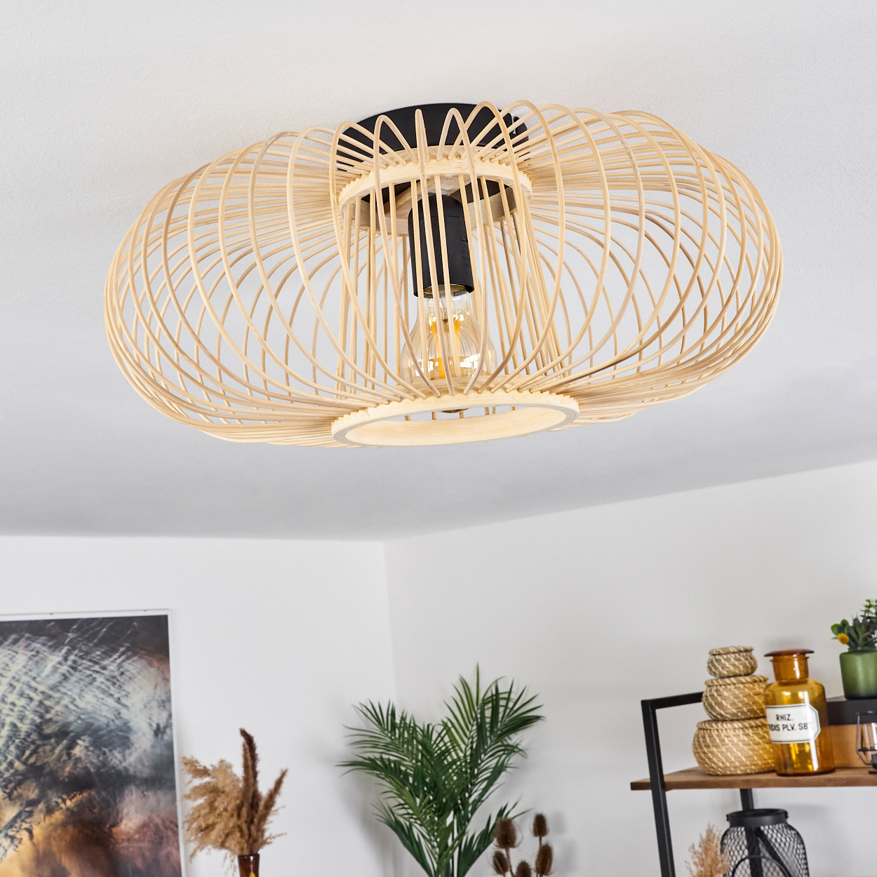 Eine gewebte, hellbraune Deckenlampe mit sichtbarer Glühbirne. Pflanzen und Dekoration sind unten zu sehen, in einem Raum mit weißen Wänden.