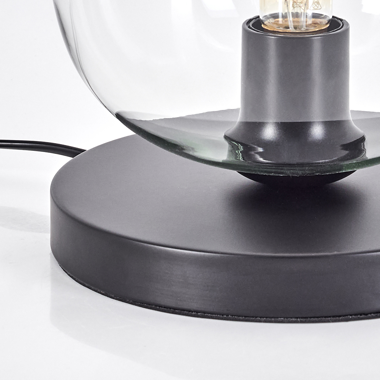 Nahaufnahme einer schwarzen Tischlampe, Glasschirm und sichtbarer Glühbirne. Der Sockel ist schwarz und rund, auf weißem Untergrund.