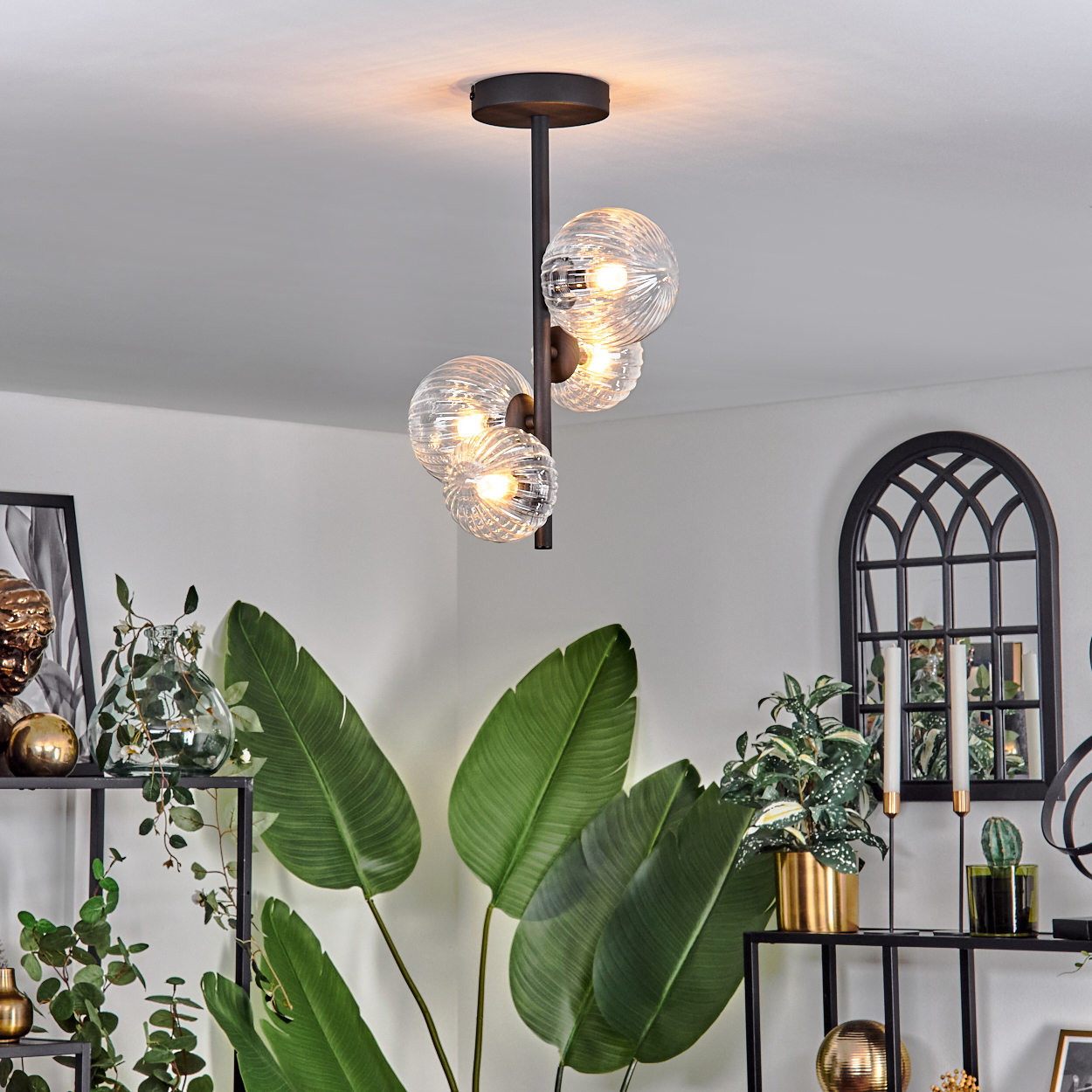 Ein modernes Interieur mit Kronleuchter. Darunter sind große grüne Blätter sichtbar. Regale mit Dekorationen wie Pflanzen und goldenen Kugeln.
