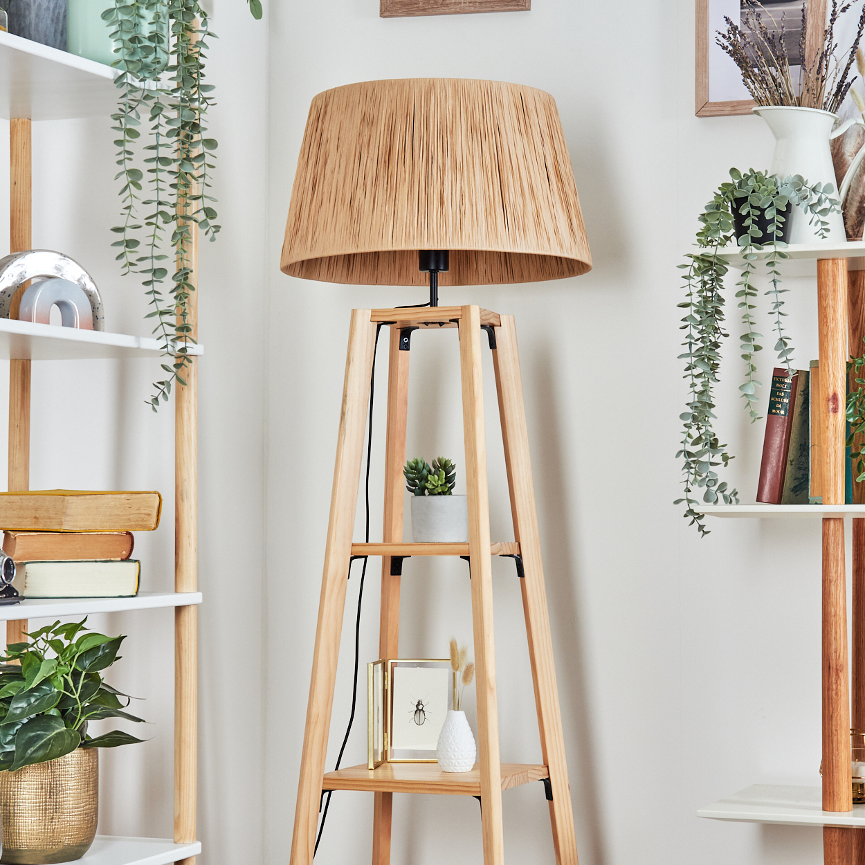 Eine Stehlampe aus Holz mit gewebtem Lampenschirm steht vor einer weißen Wand. Regale auf beiden Seiten halten Pflanzen und Bücher.