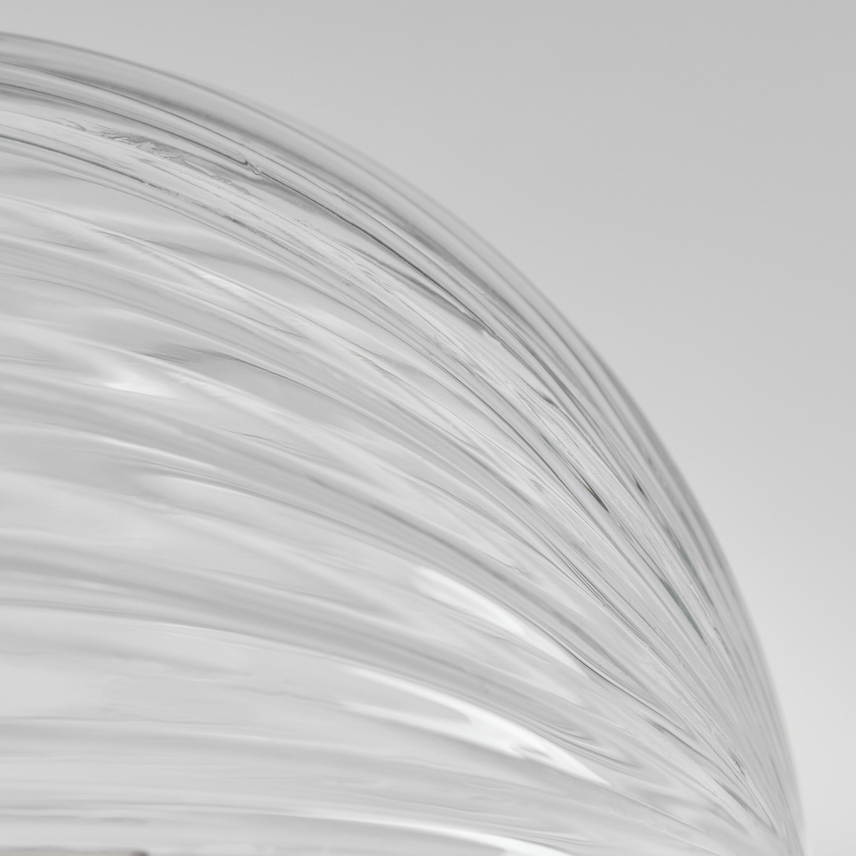 Nahaufnahme eines transparenten, gebogenen Glasobjekts. Parallele Linien erzeugen eine strukturierte Oberfläche vor grauem Hintergrund, abstrakt.